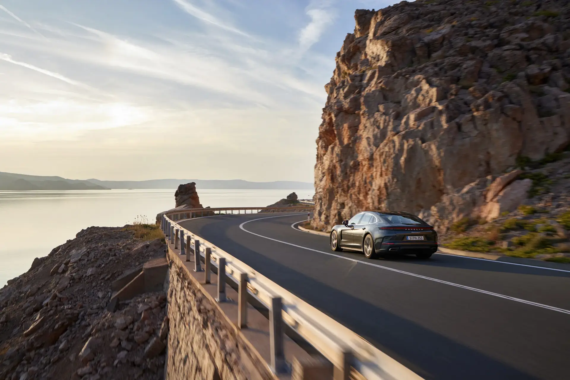 En mørk Porsche Panamera kører langs en snoet kystvej ved klipper og vand i aftensol.
