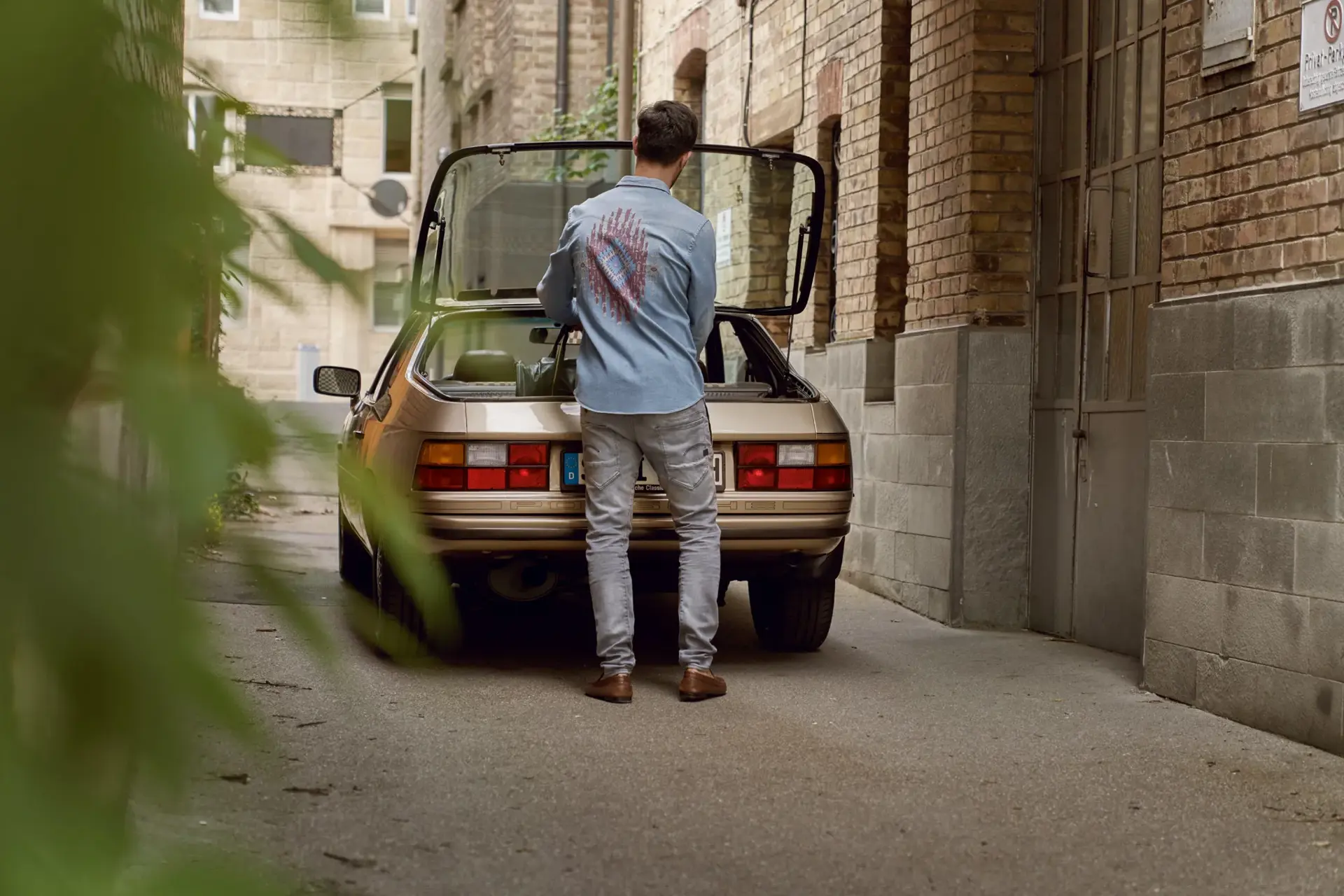 En mand står i en smal gård og pakker bagagerummet på en gylden klassisk Porsche.