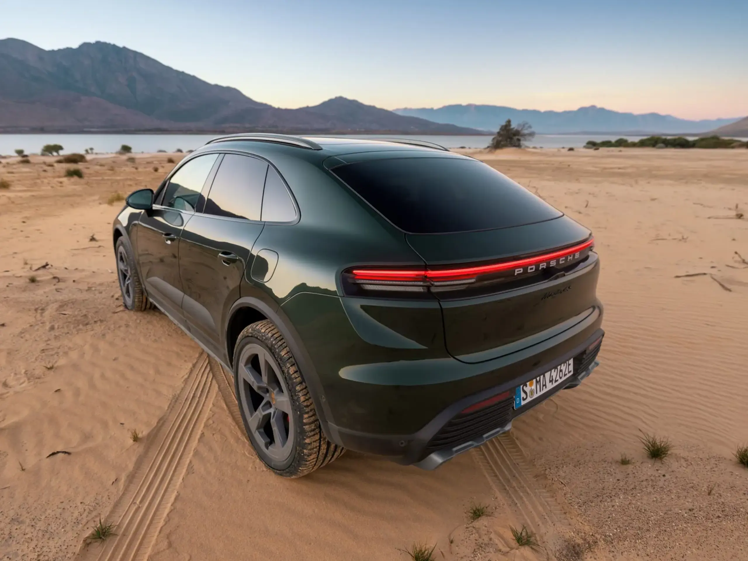 Mørkegrøn Porsche Macan 4S set skråt bagfra, parkeret i sandet ved en sø med bjerge i baggrunden.