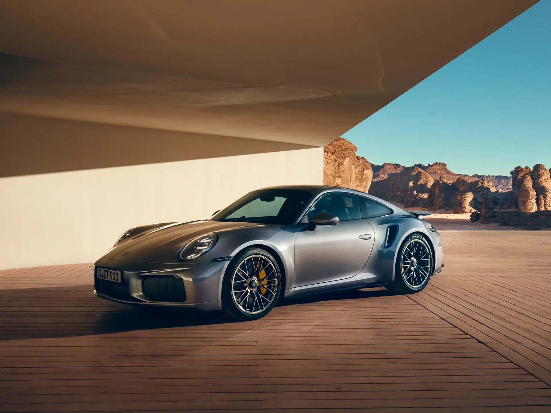 En sølvgrå Porsche 911 Turbo S holder parkeret under et moderne halvtag med en solbeskinnet ørken og klippeformationer i baggrunden.