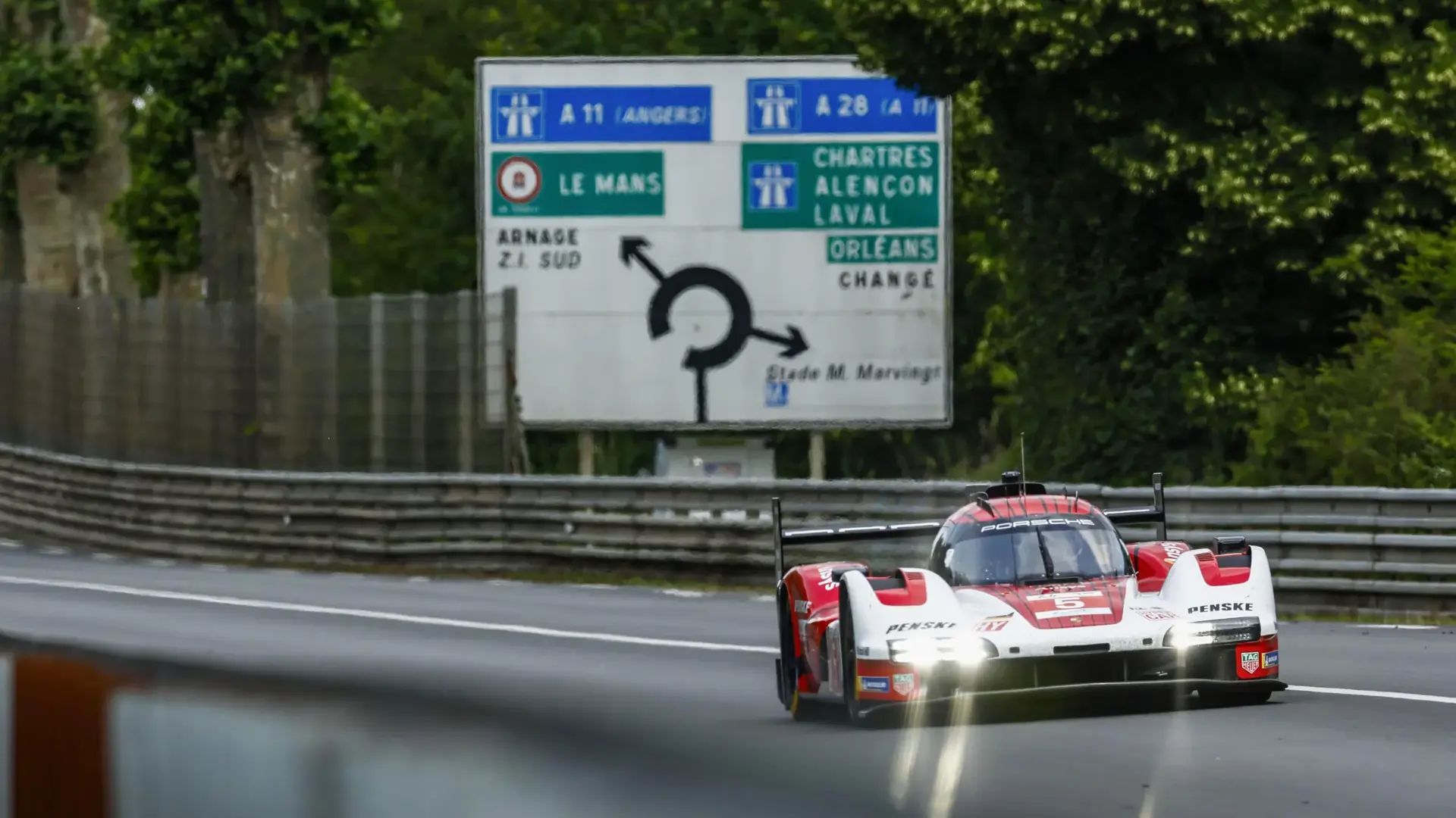 En rød og hvid Porsche Le Mans-racer kører i høj fart på banen foran et stort fransk motorvejsskilt.