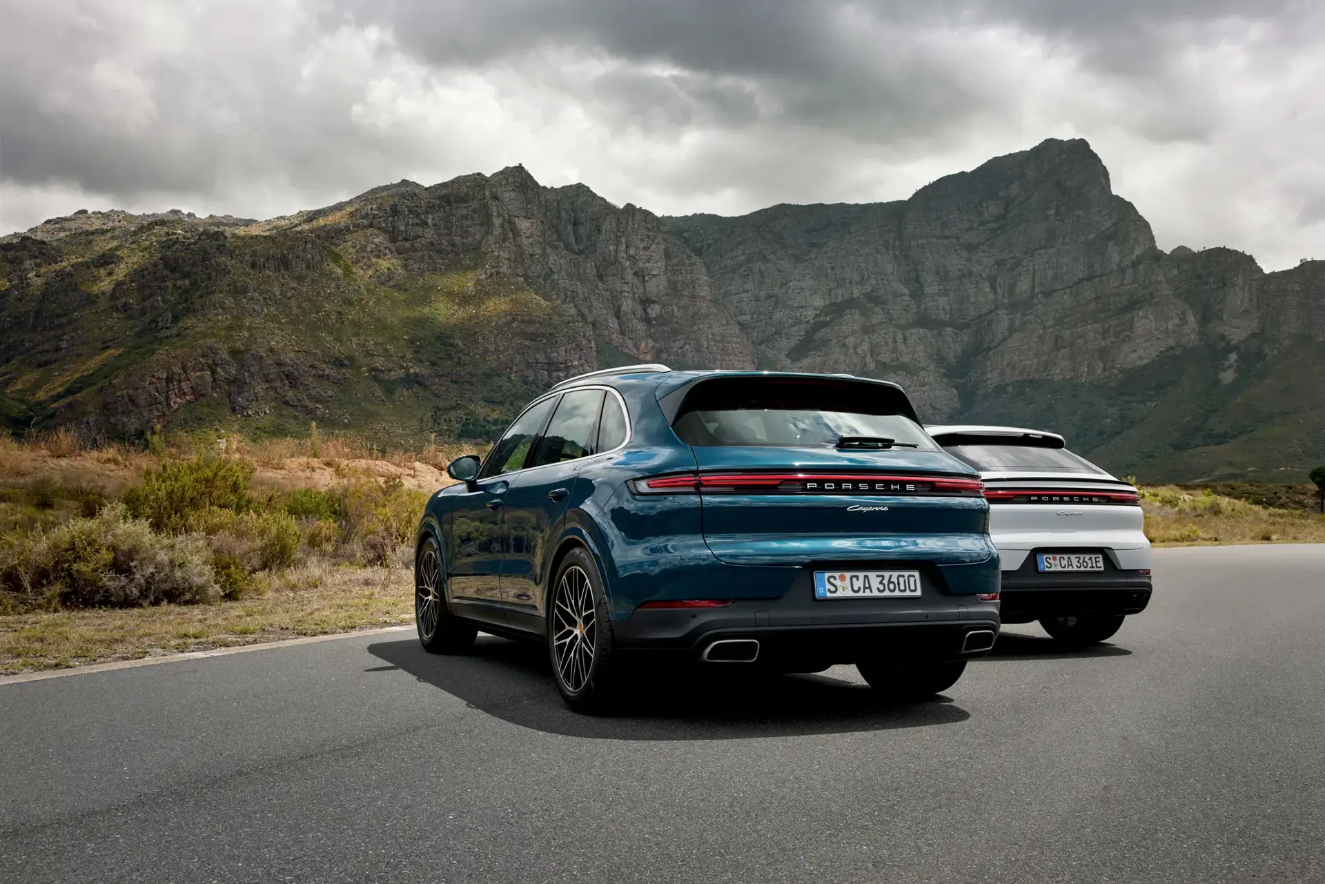 To Porsche Cayenne SUV’er holder parkeret på en vej foran dramatiske bjerglandskaber under en overskyet himmel.