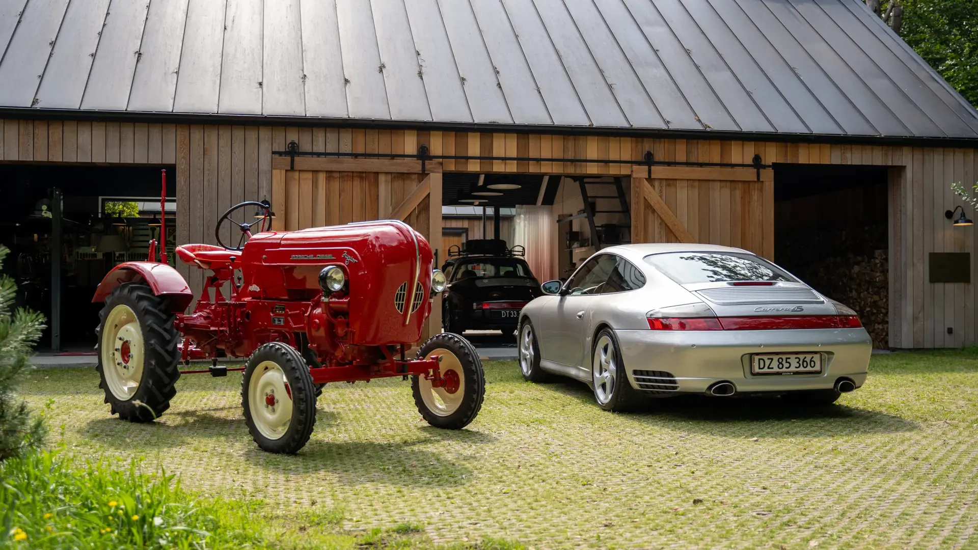 En rød vintage traktor og en sølv Porsche 911 står parkeret foran en moderne trægarage med åbne porte.