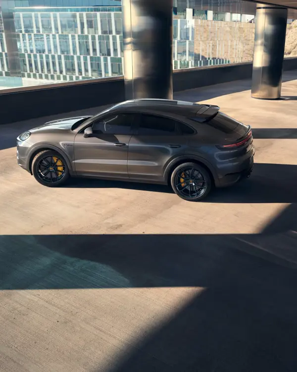 En grå Porsche Cayenne Turbo E-Hybrid Coupé med gule bremsekalibre holder parkeret i skyggen ved en moderne glasbygning.
