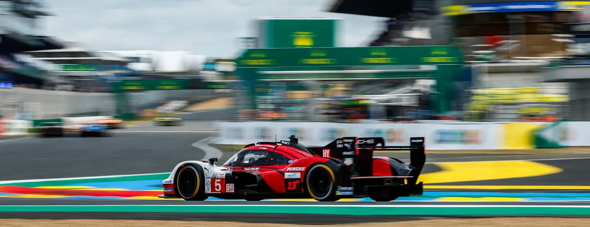 En rød og sort Porsche-hypercar i høj fart på Le Mans-banen med slørede tribuner i baggrunden.