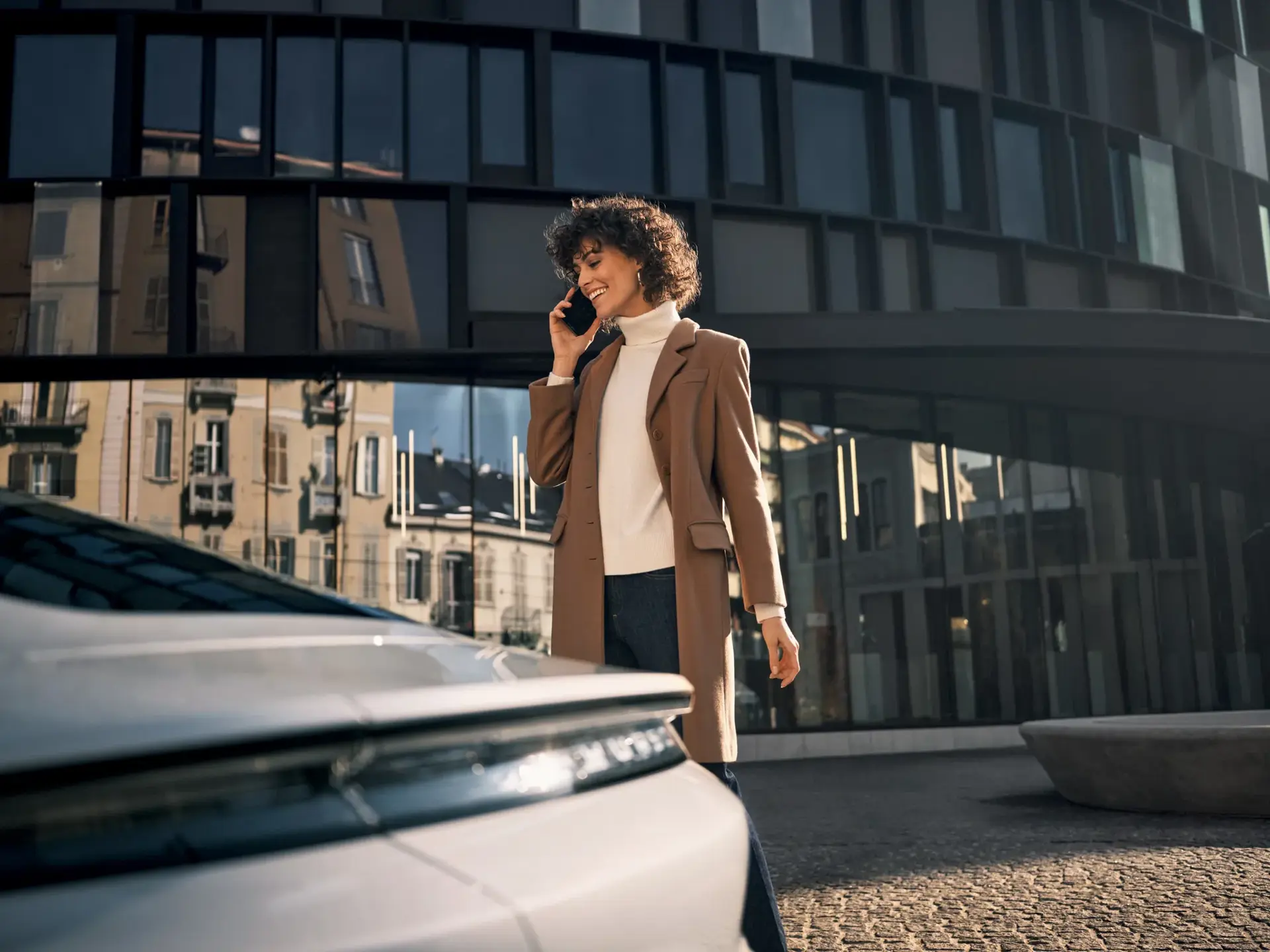 En smilende person går talende i telefon ved siden af en hvid Porsche foran en moderne glasbygning i byen.