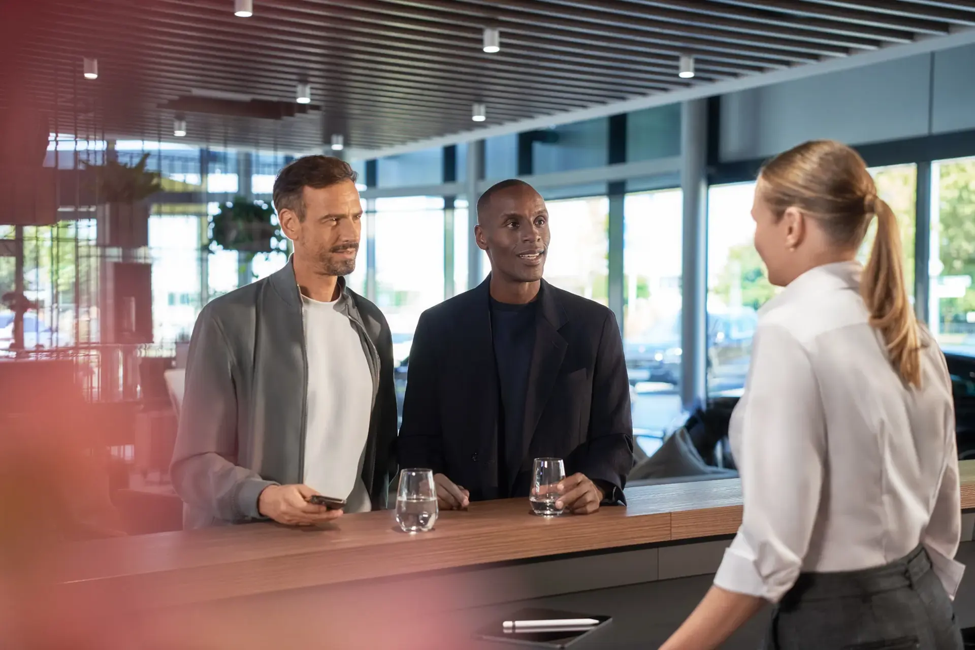 To mænd står ved en skranke og taler med en kvinde i en moderne, lys reception.