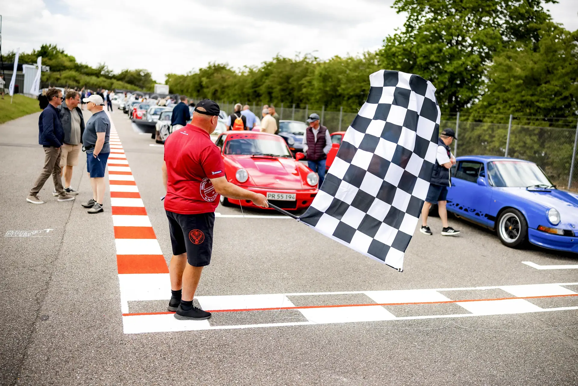 En mand svinger et ternet racerflag ved startlinjen foran en række klassiske Porsche-biler og tilskuere.