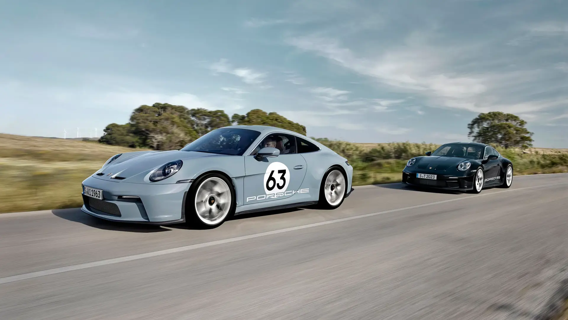To Porsche 911-sportsvogne kører hurtigt side om side på en landevej i åbent landskab.