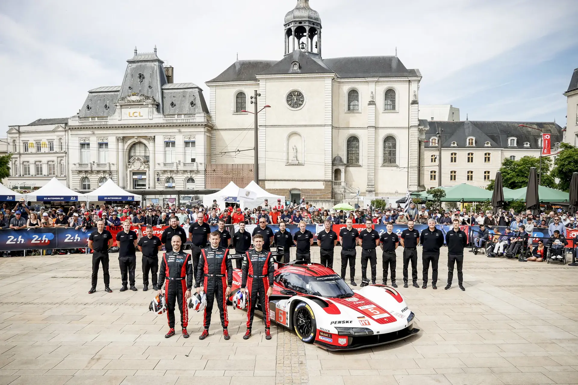 Racerteam poserer foran en rød-hvid Porsche LMDh-racerbil på en plads med historiske bygninger og publikum i baggrunden.