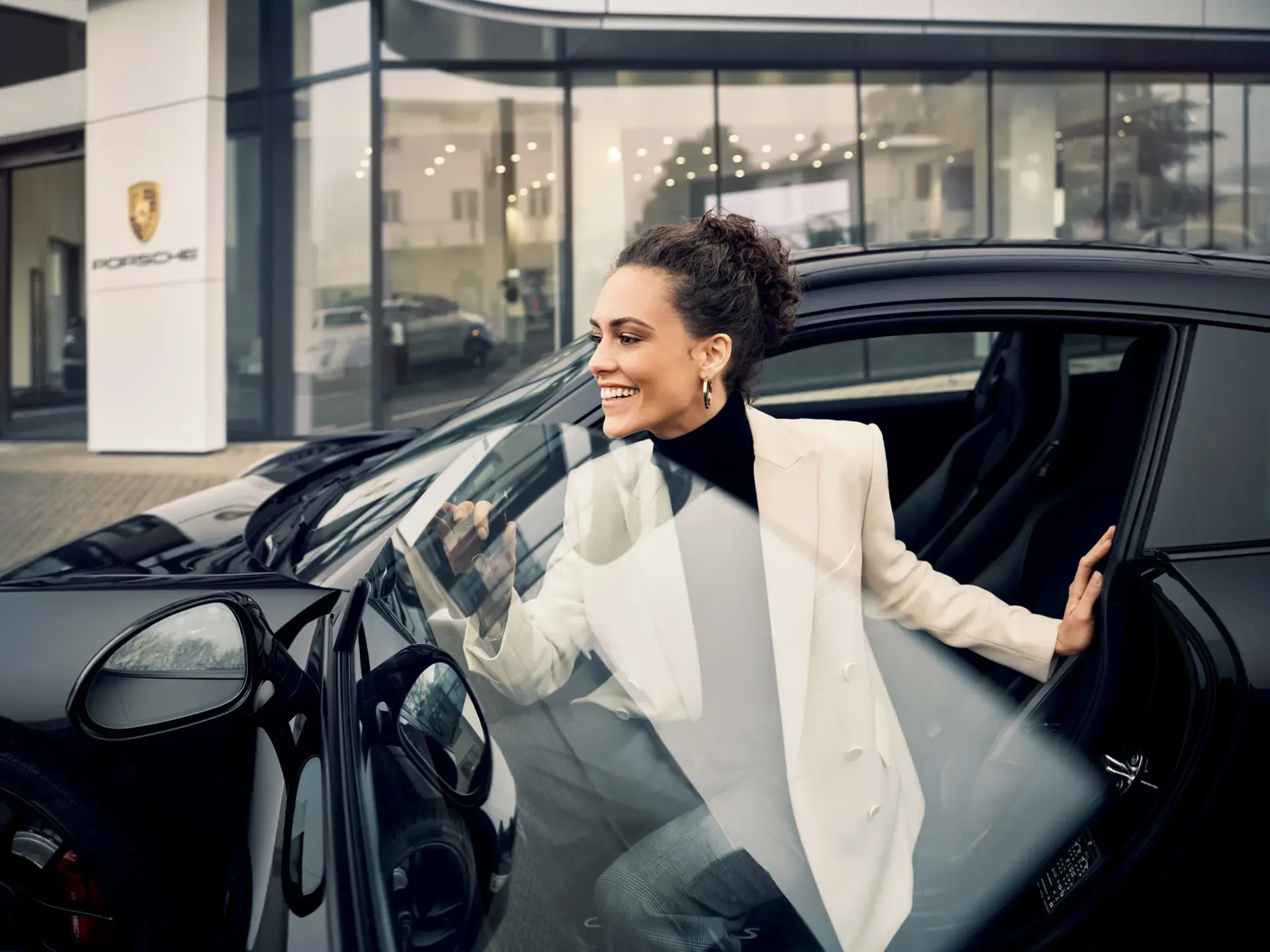 Kvinde i elegant tøj stiger smilende ind i en sort Porsche foran et Porsche-center.