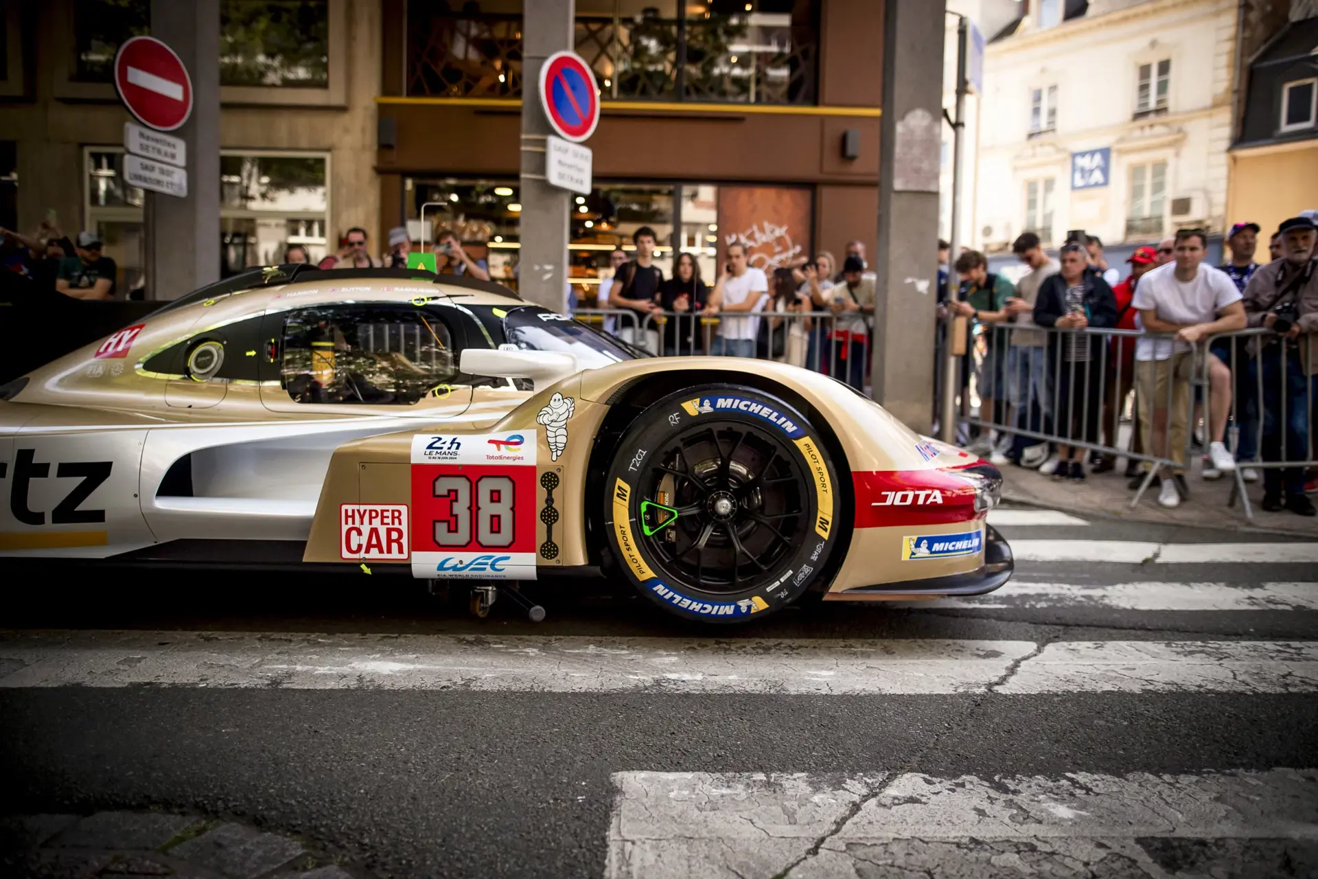 En guldfarvet hypercar med startnummer 38 passerer en fodgængerovergang i byen foran tilskuere bag afspærringer.