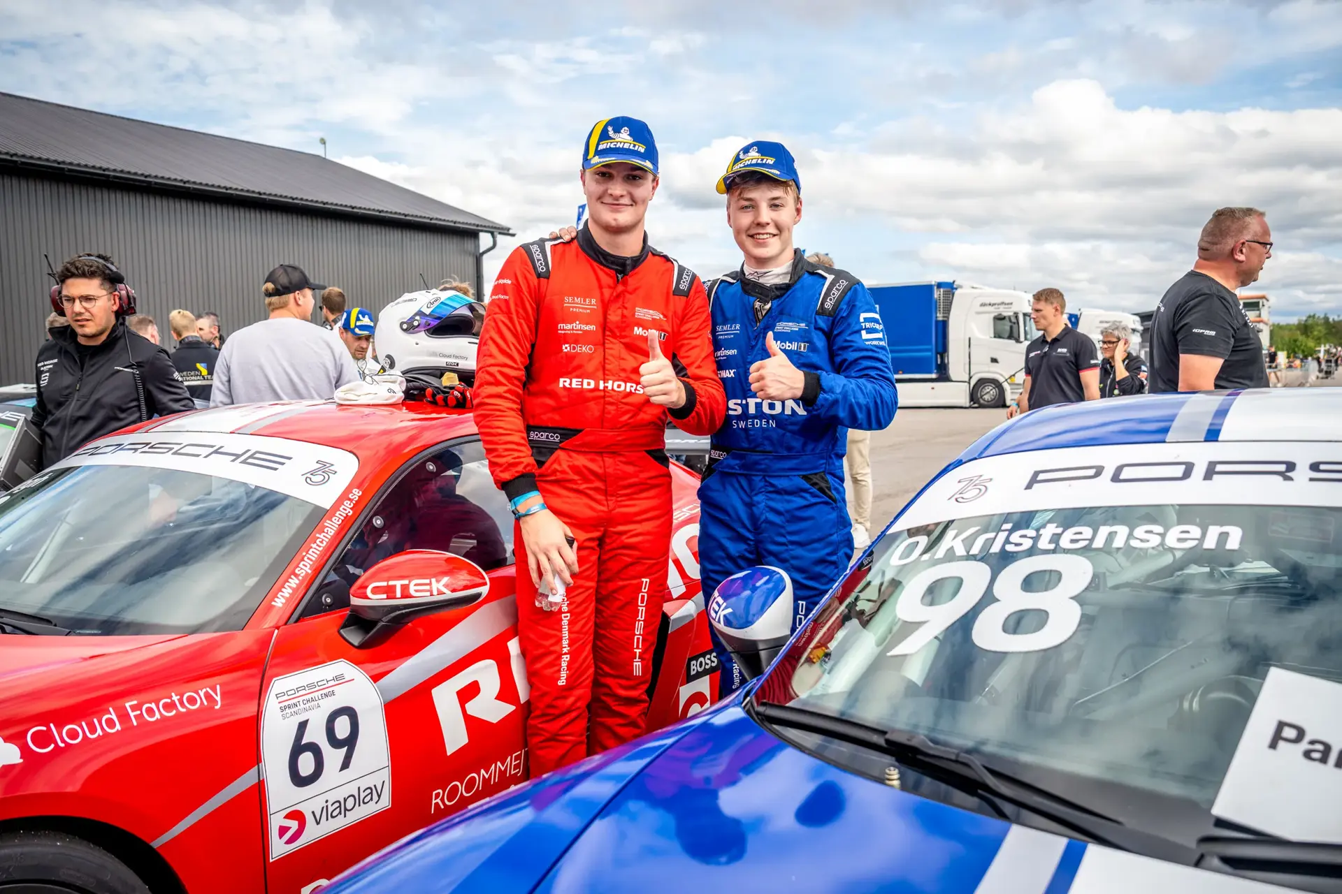 To racerkørere i rødt og blåt køredragt står mellem deres Porsche-løbsbiler i pitområdet og giver thumbs up før et løb.