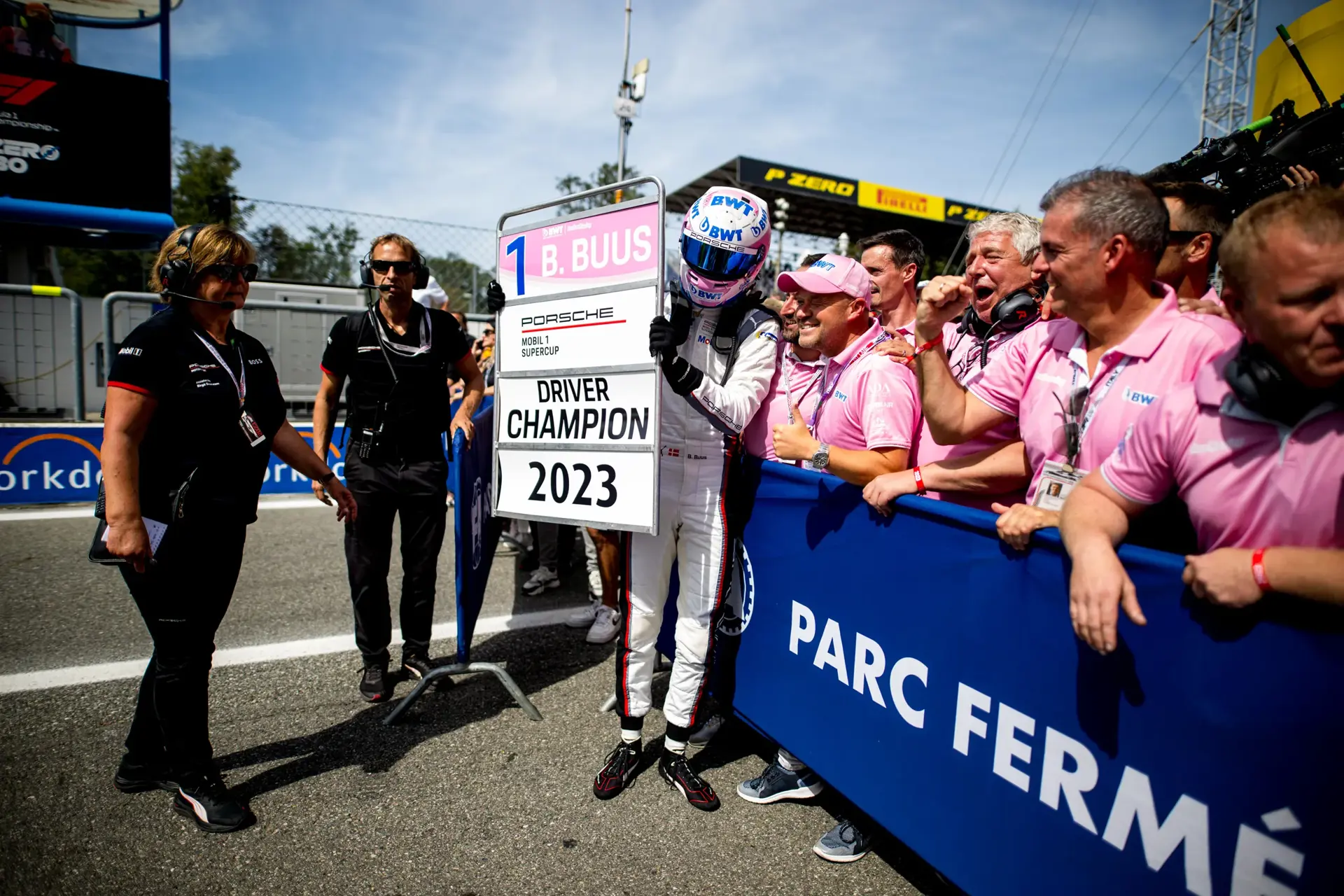 Racerkører i køredragt fejrer et mesterskab ved Monza og holder et stort skilt med teksten "Driver Champion 2023" foran jubelende teammedlemmer.