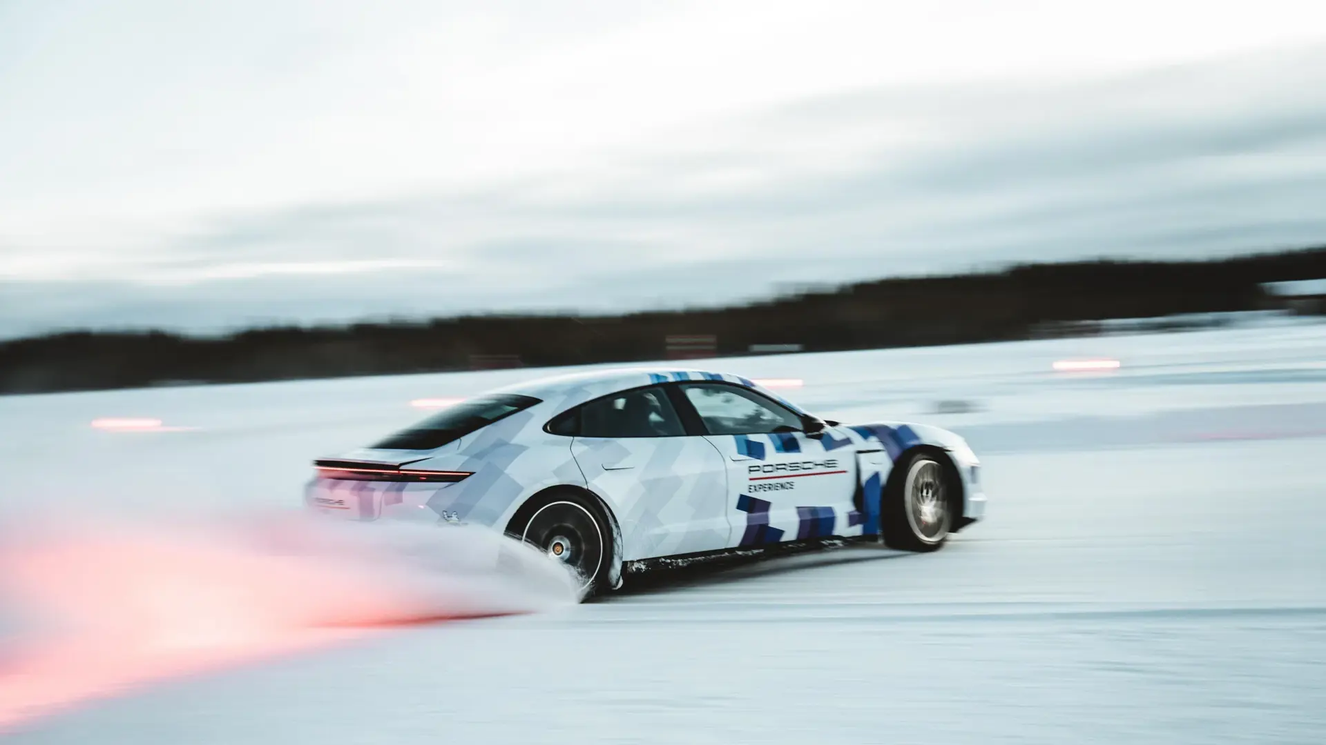 En hvid og blå Porsche sportsvogn drifter hurtigt hen over en snedækket bane med sne og lysstriber bag sig.