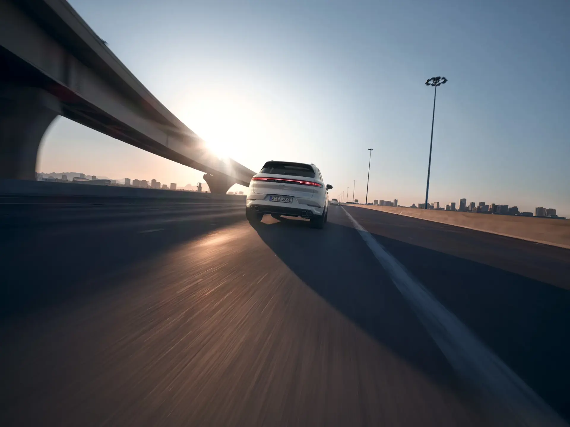 En hvid Porsche Cayenne Turbo e-Hybrid ses bagfra, mens den kører på en motorvej mod en by i solnedgang.