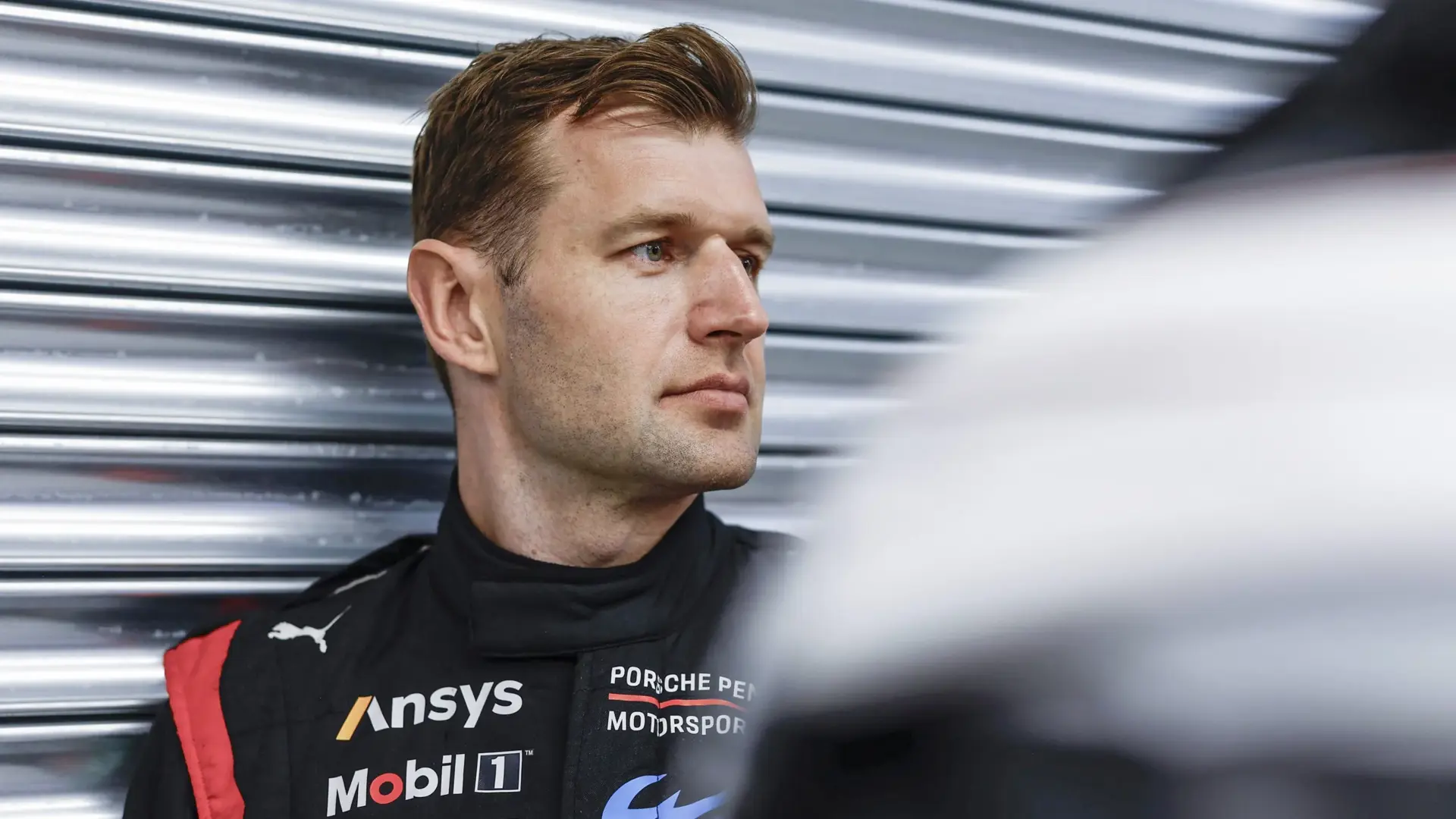 Racer i sort Porsche Motorsport-kørerdragt står foran en metalport i pitområdet.