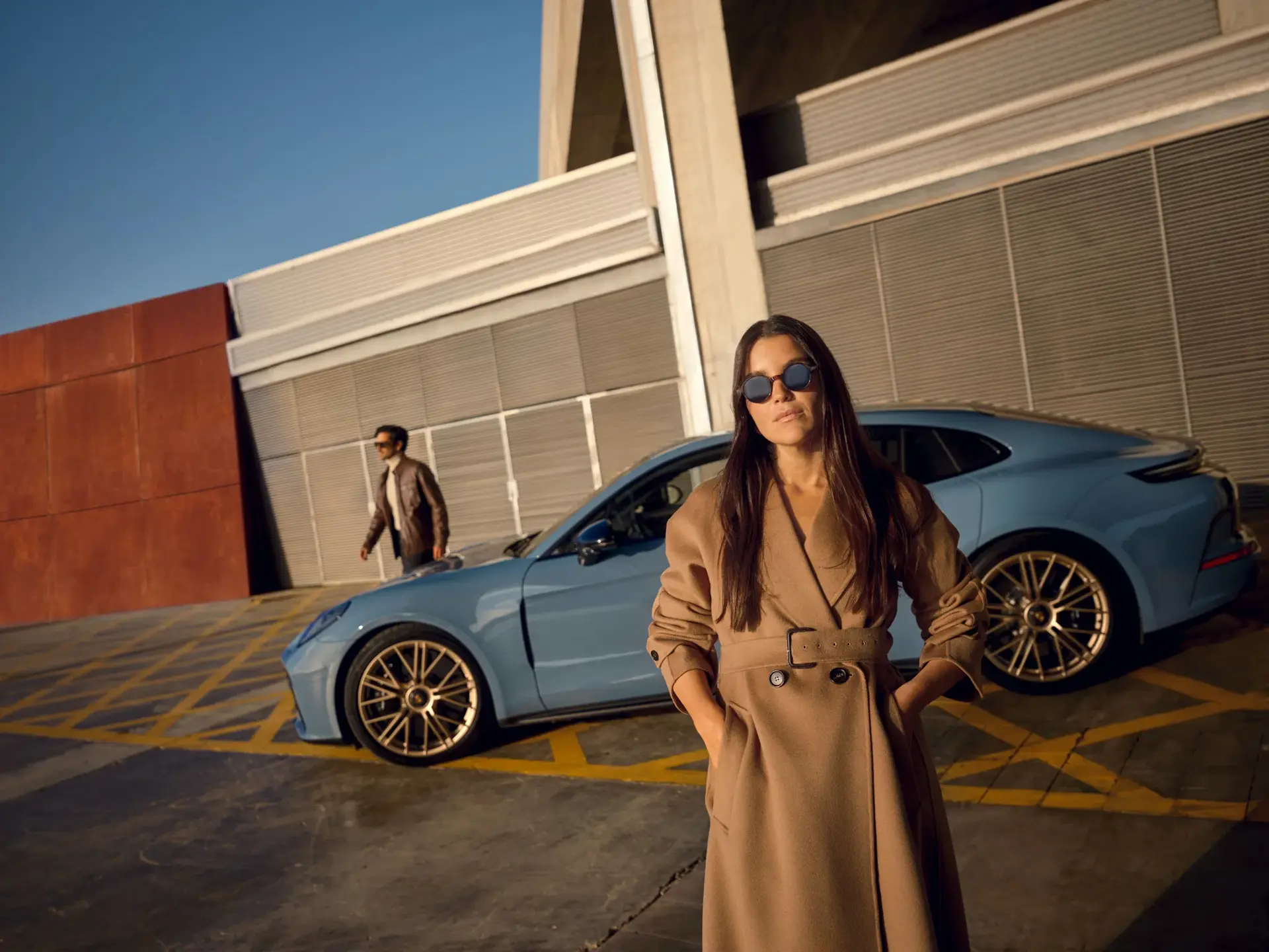 To personer poserer foran en blå Porsche Panamera i et moderne, industrielt parkeringsmiljø.