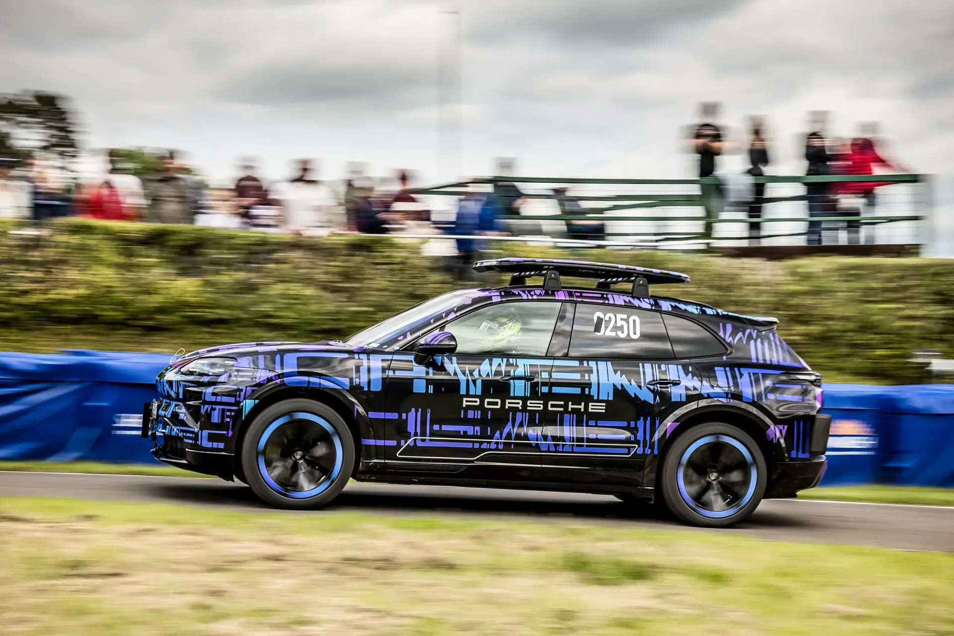 En sort Porsche SUV med lilla og blå camouflagelak kører hurtigt forbi tilskuere langs en racerbane.