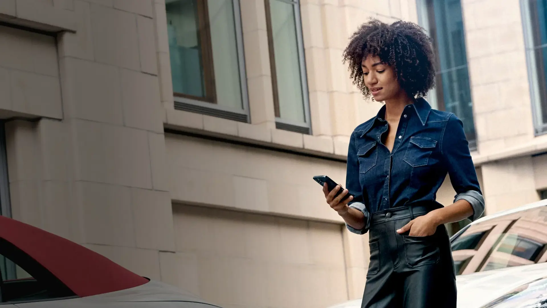 En kvinde går langs en bygning og kigger på sin smartphone ved siden af en parkeret bil.