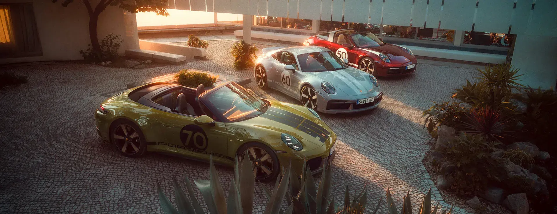 Tre Porsche sportsvogne i heritage-design holder parkeret i en solbeskinnet, moderne gårdhave omgivet af planter.