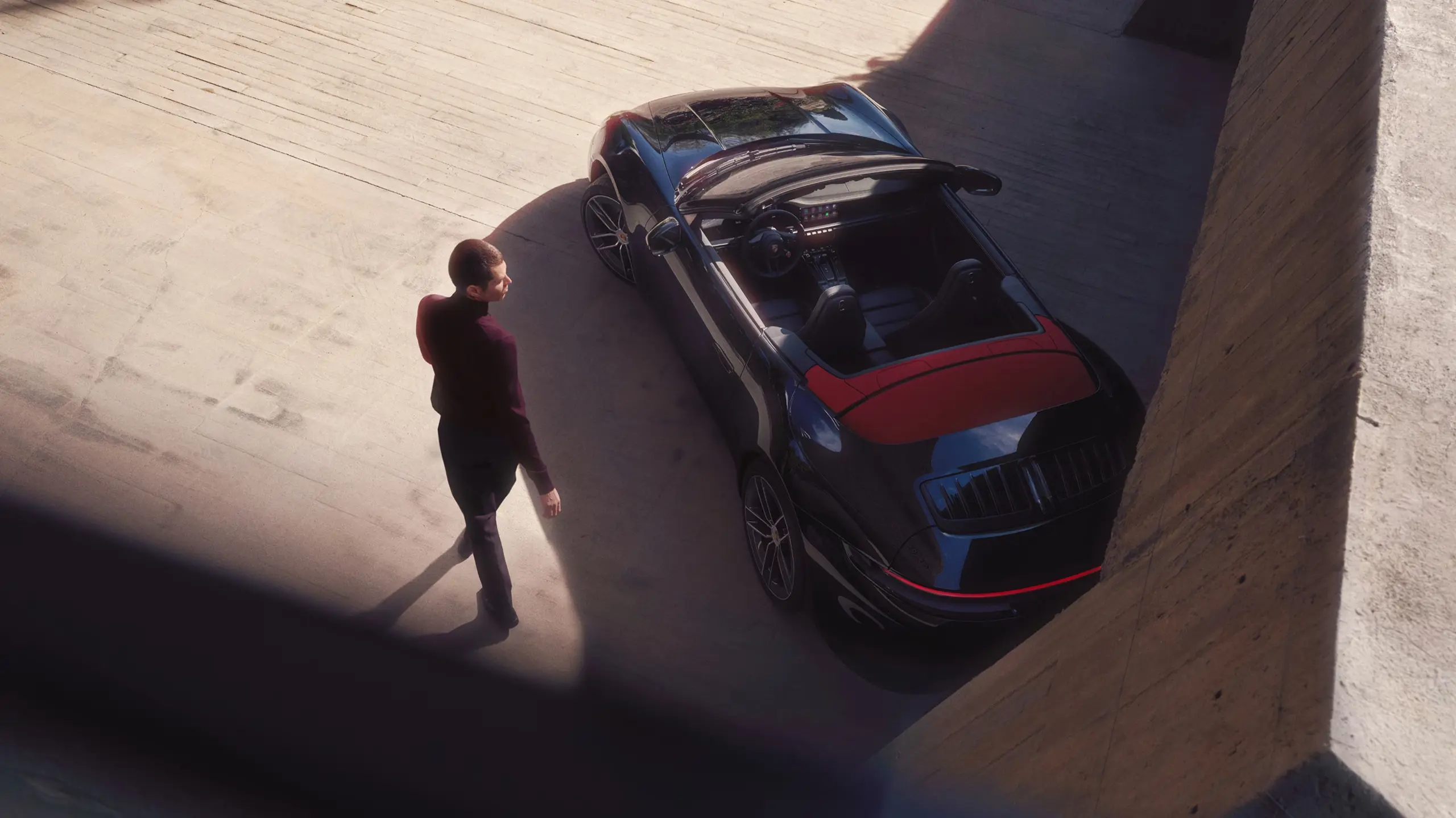 En tatoveret mand i vinrød sweater sidder i førersædet på en Porsche 911 Cabriolet og kigger op mod kameraet.