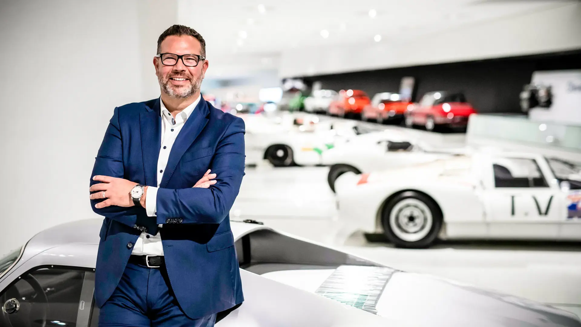 Mand i blå jakkesæt står med korslagte arme foran klassiske sportsvogne i et lyst bilmuseum.
