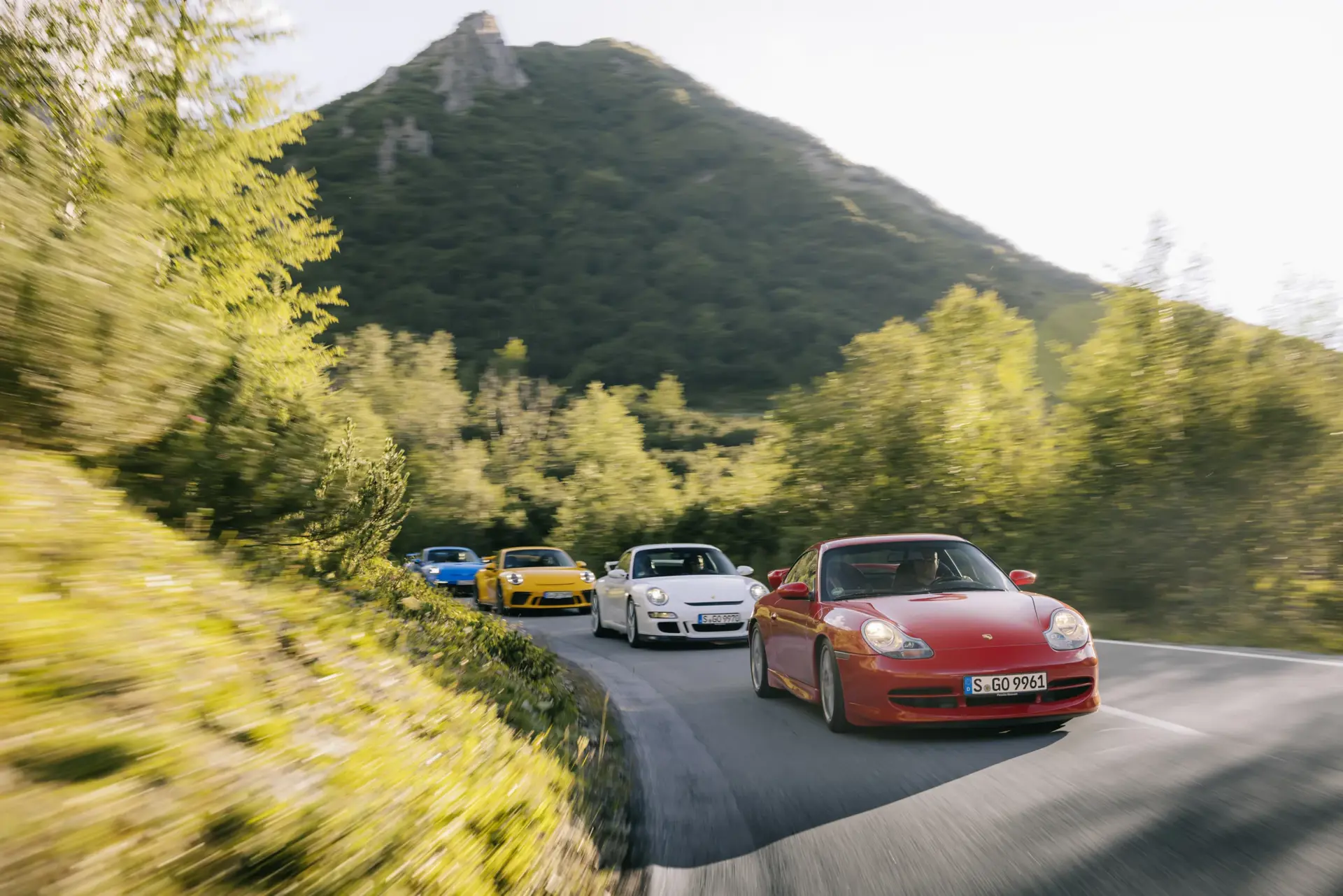 Flere farverige Porsche 911 sportsbiler kører hurtigt gennem en snoet bjergvej omgivet af grøn natur.