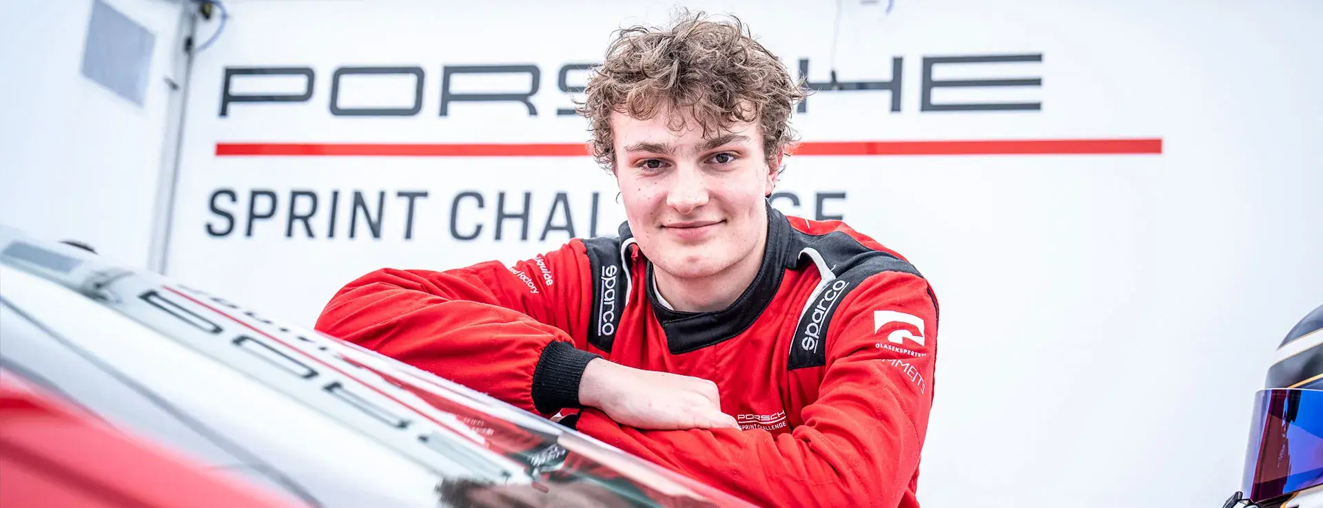 Racer i rød køredragt læner sig mod en Porsche-racerbil foran et “Porsche Sprint Challenge”-banner.