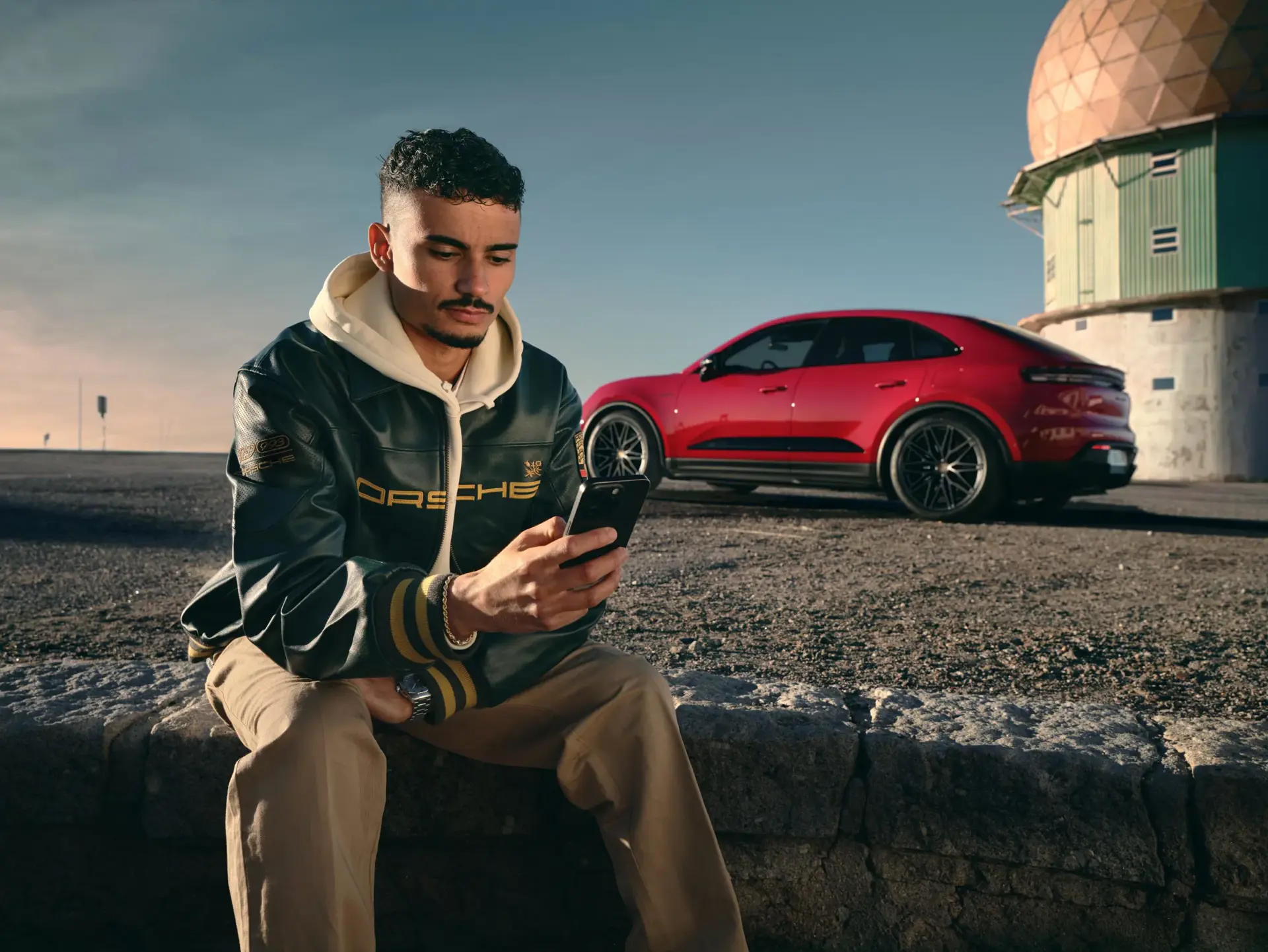 En mand sidder og bruger sin smartphone foran en rød Porsche Macan parkeret i et øde landskab.