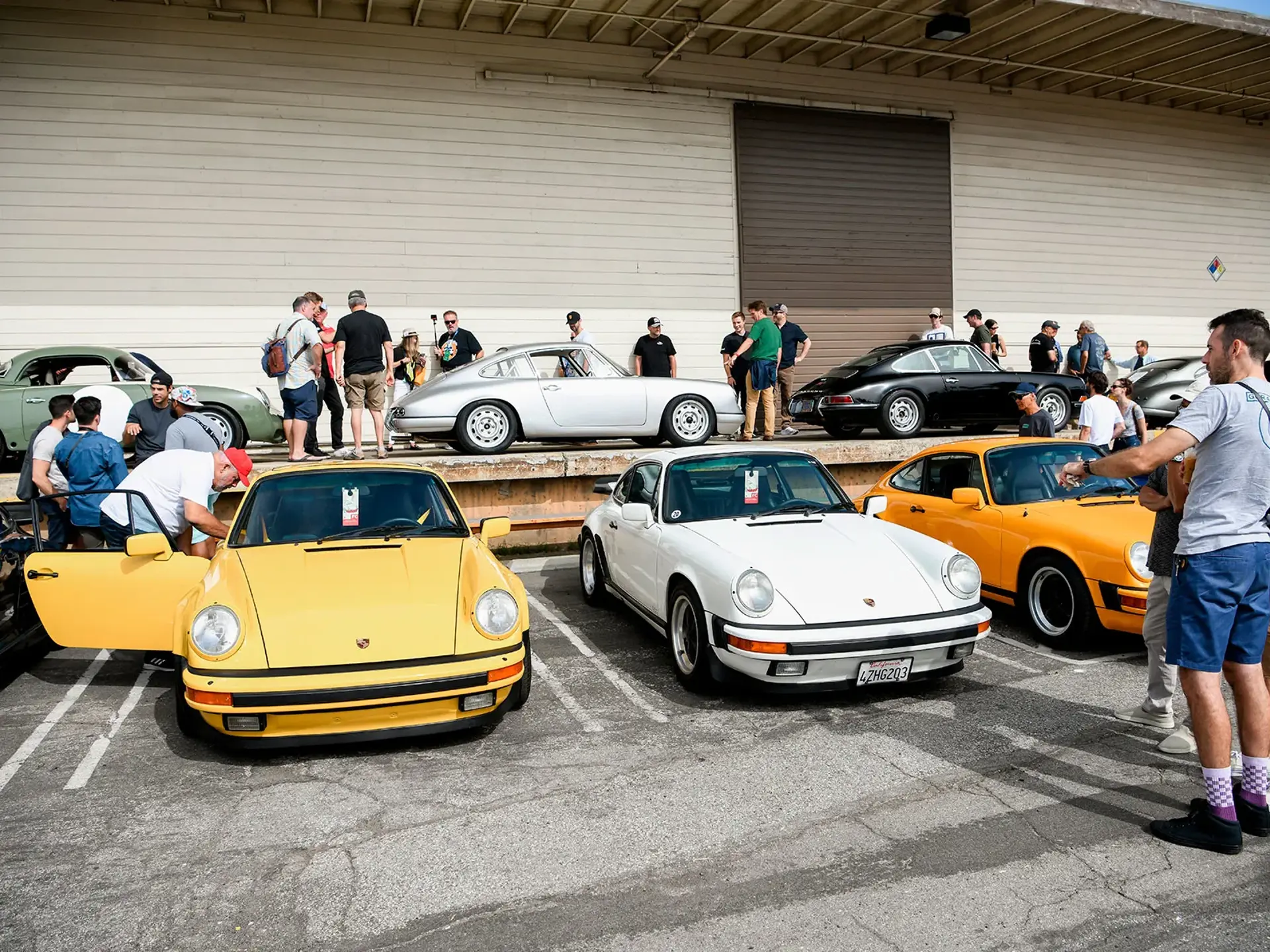 En gruppe mennesker samles omkring en række klassiske Porsche 911 sportsvogne parkeret foran en industribygning.