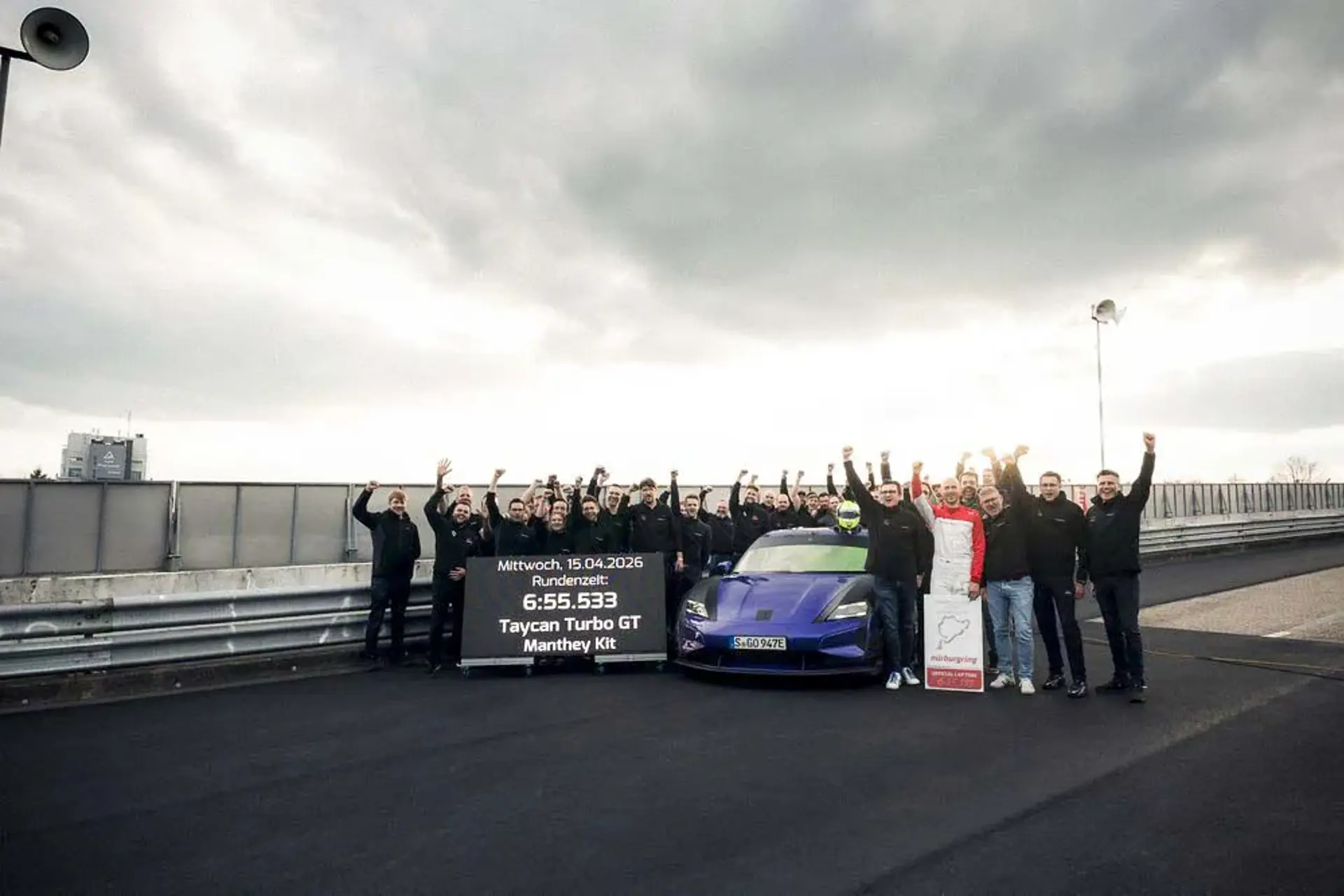 Et hold mennesker fejrer ved siden af en blå Porsche Taycan Turbo GT på en racerbane med et skilt, der viser en ny omgangsrekord.