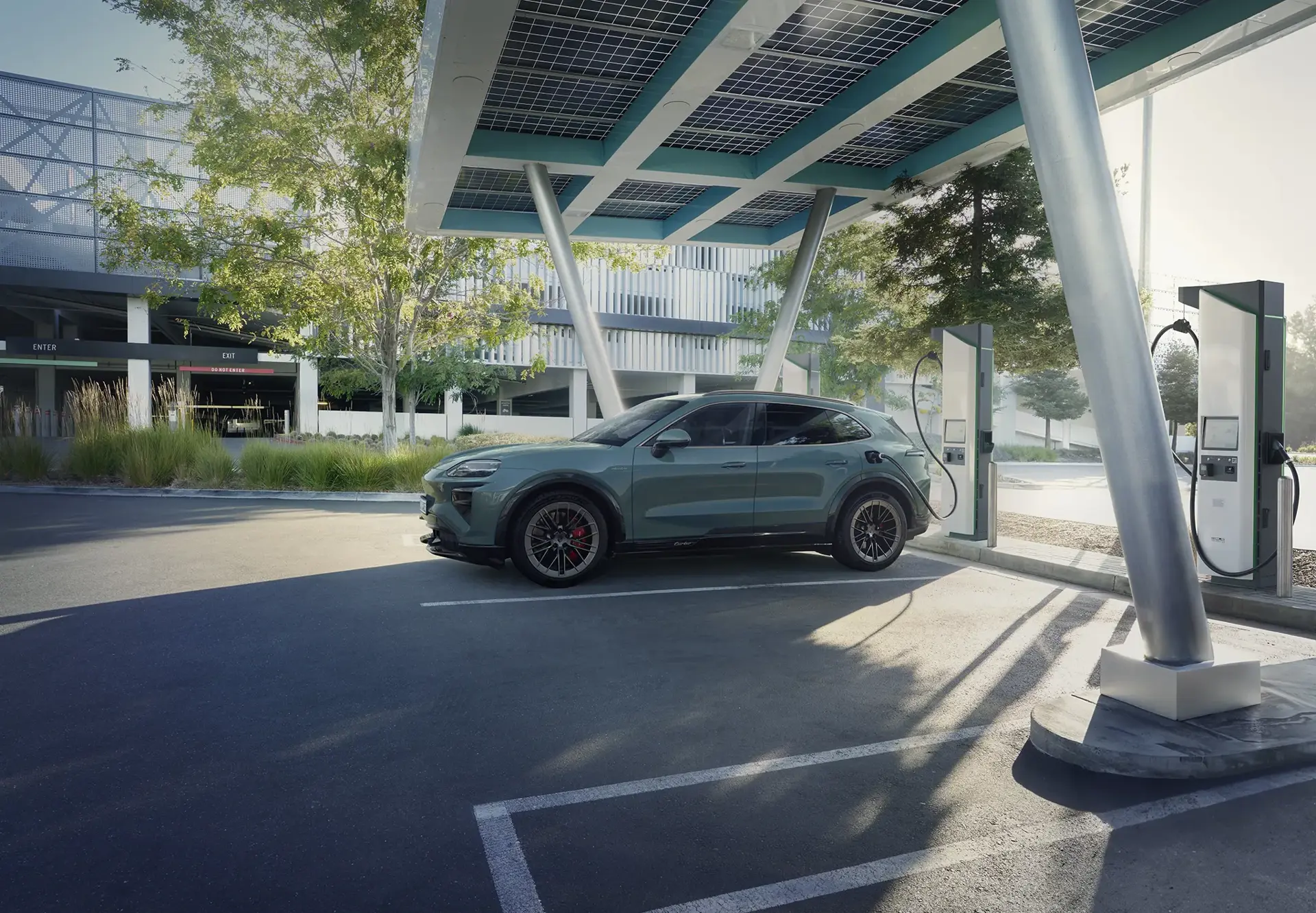 En grøn Porsche Cayenne Turbo E-Hybrid står til opladning ved en moderne ladestation under et solcelletag i et parkeringsanlæg.