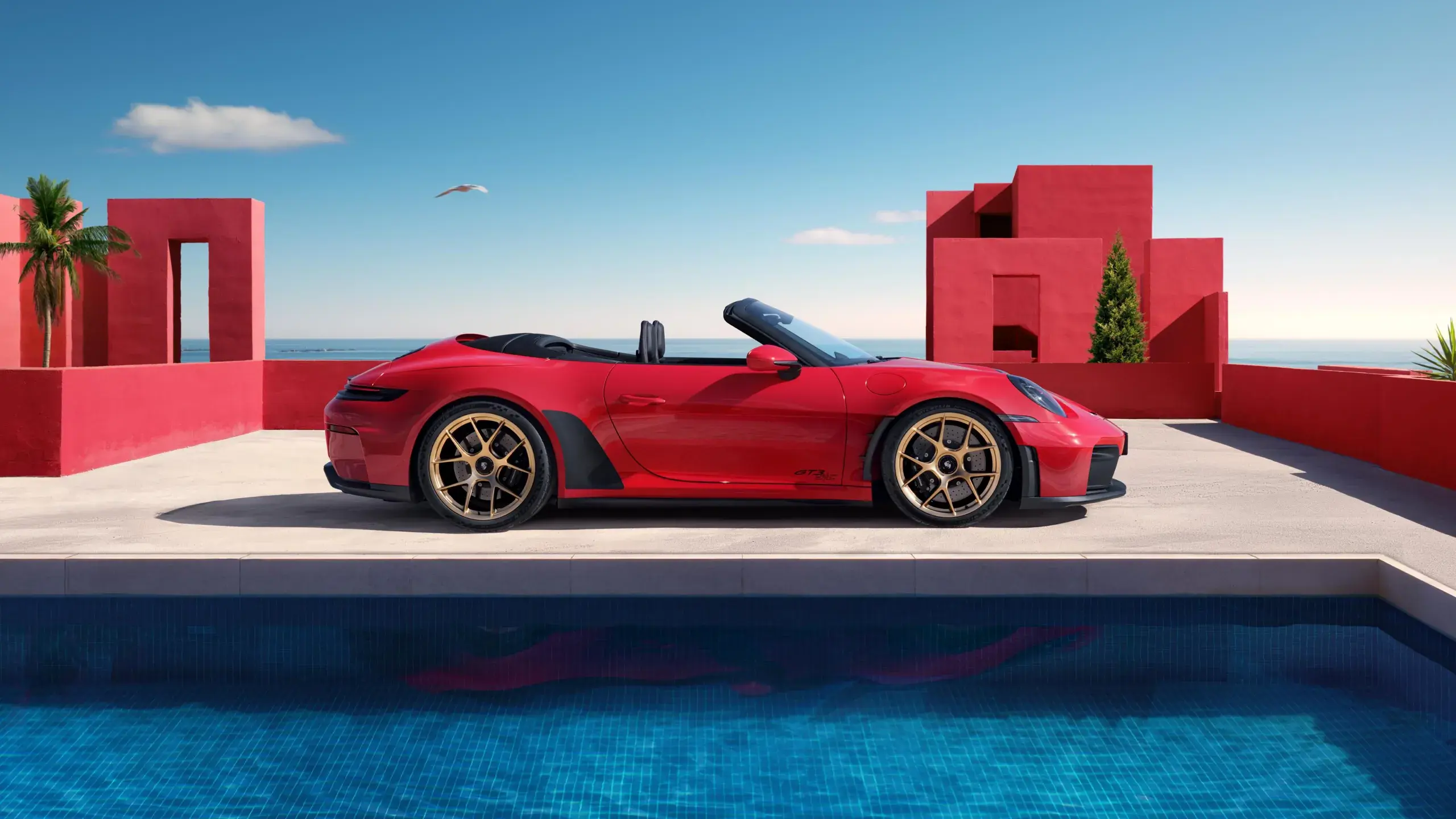 En rød Porsche 911 cabriolet holder parkeret ved en pool foran moderne røde bygninger med havet i baggrunden.