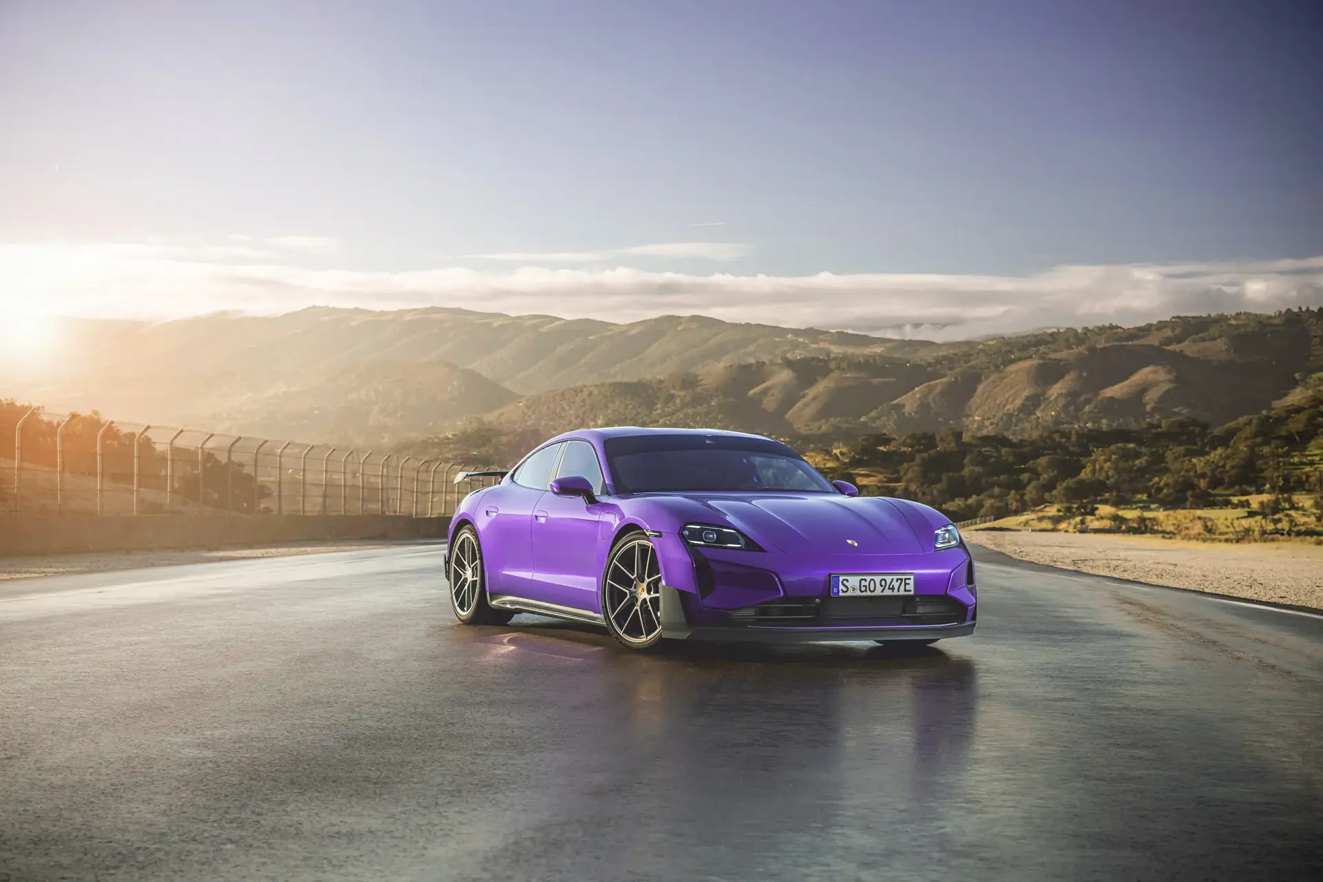 Lilla Porsche Taycan Turbo GT står parkeret på en våd racerbane med bjerge i baggrunden ved solnedgang.
