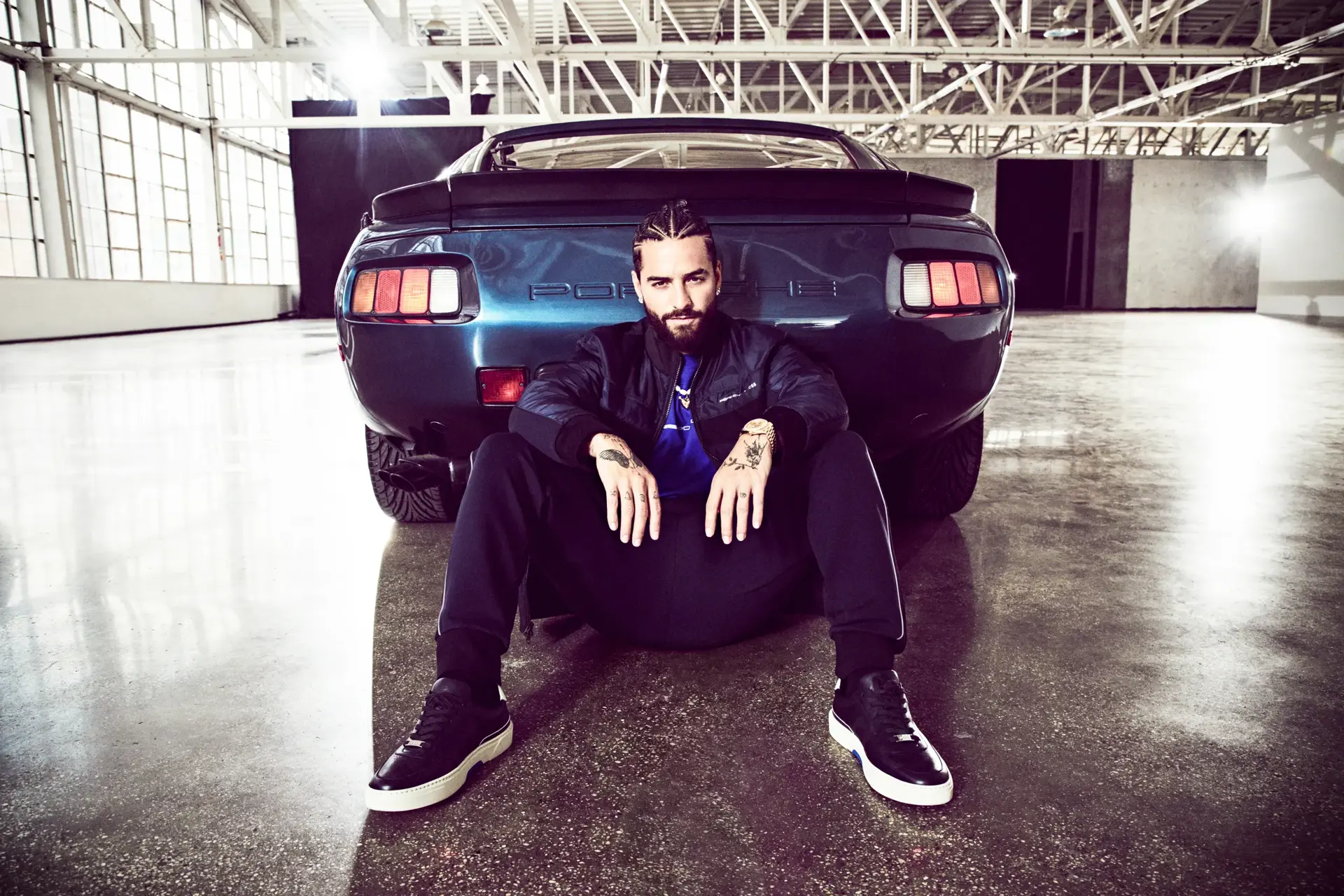 Person i sporty BOSS-outfit sidder afslappet op ad bagenden af en blå Porsche i en lys industribygning.