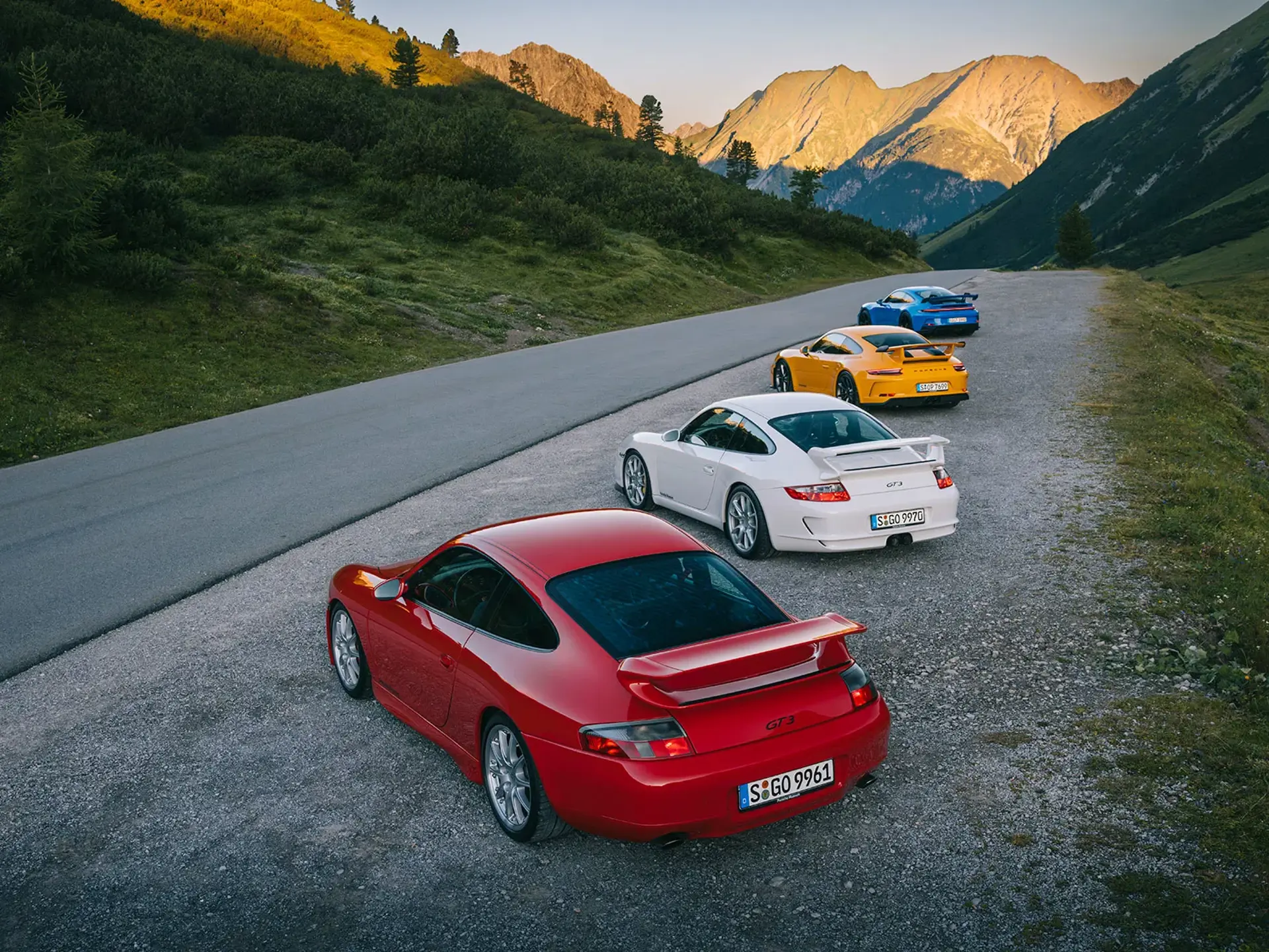 Fem farverige Porsche 911 GT3 sportsvogne holder parkeret på en bjergvej i solnedgangens lys.