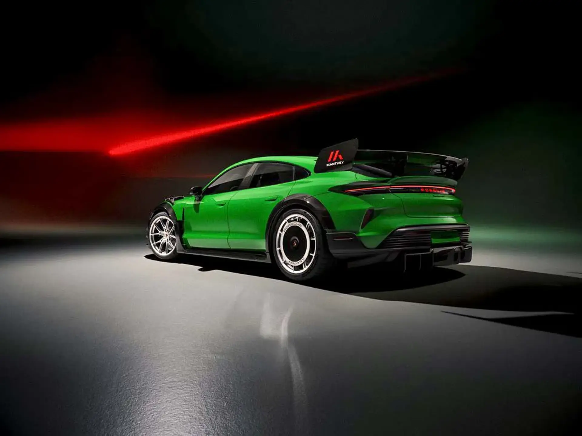 Grøn Porsche Taycan Manthey med stor hækspoiler set skråt bagfra i et mørkt studie med rød lyseffekt.
