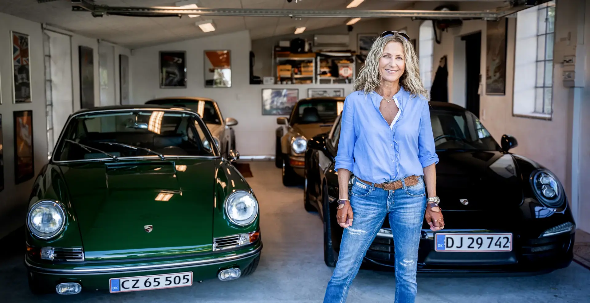 Smilende kvinde står i en garage omgivet af flere klassiske Porsche-sportsvogne.