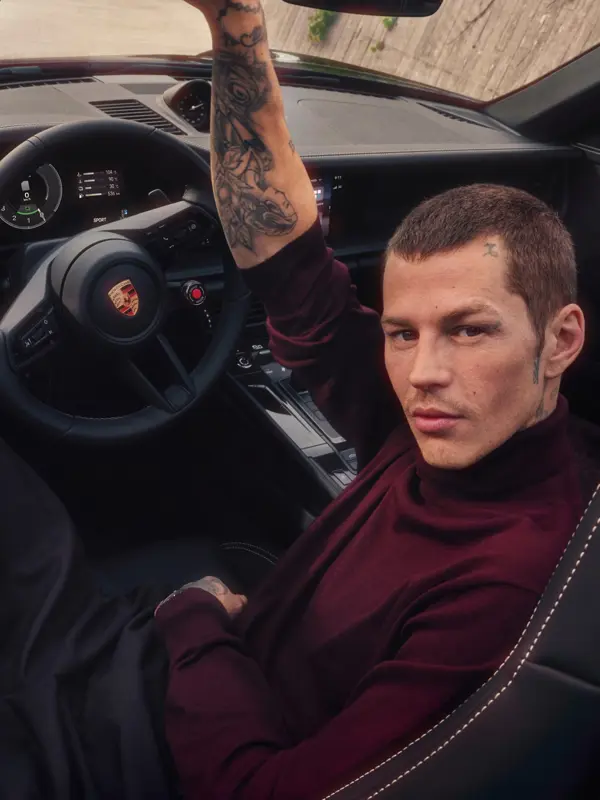En tatoveret mand i vinrød sweater sidder i førersædet på en Porsche 911 Cabriolet og kigger op mod kameraet.
