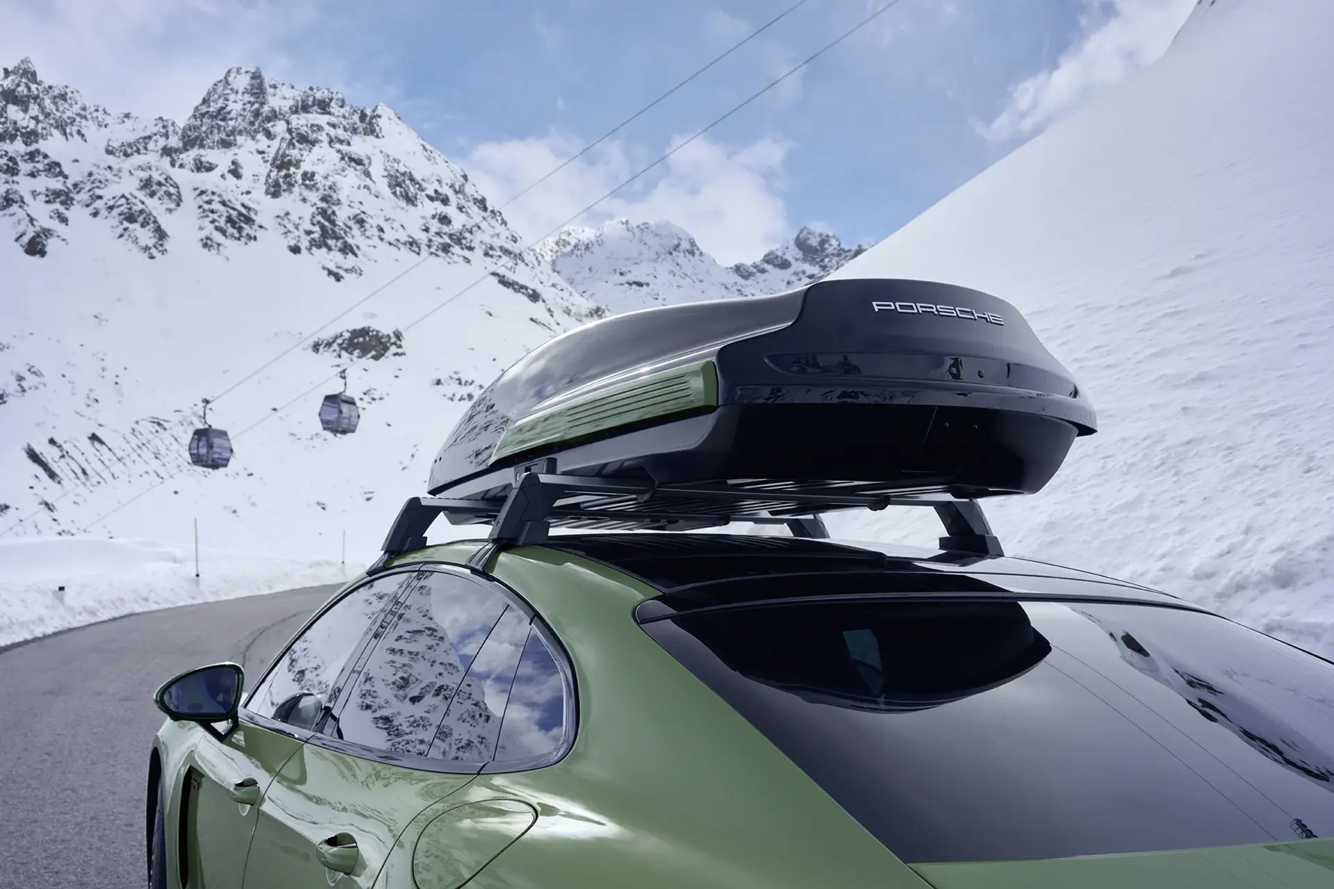En grøn Porsche Panamera med tagboks kører i et sneklædt bjerglandskab med skilifter i baggrunden.