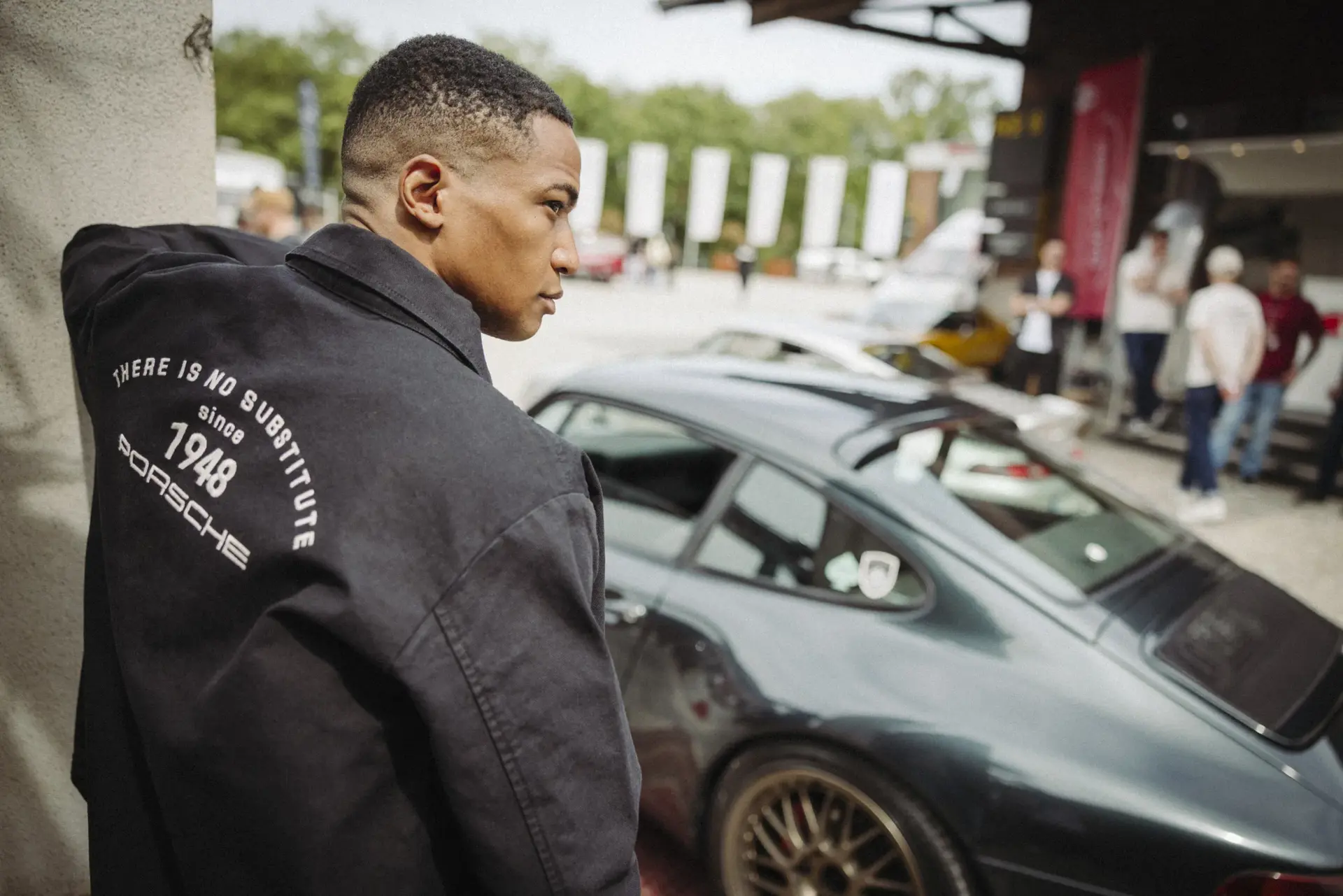 Mand i Porsche-jakke står ved siden af en sort Porsche sportsvogn til et udendørs bilarrangement.