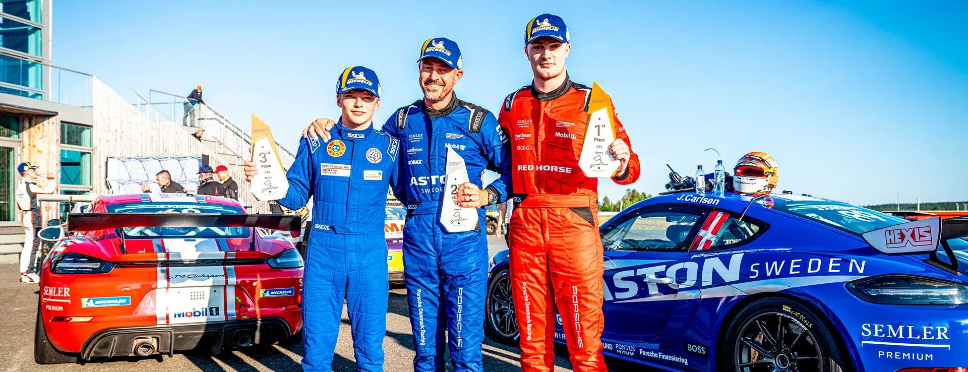 Tre racerkørere i køredragter står mellem to Porsche-racerbiler og holder pokaler efter et løb på banen.