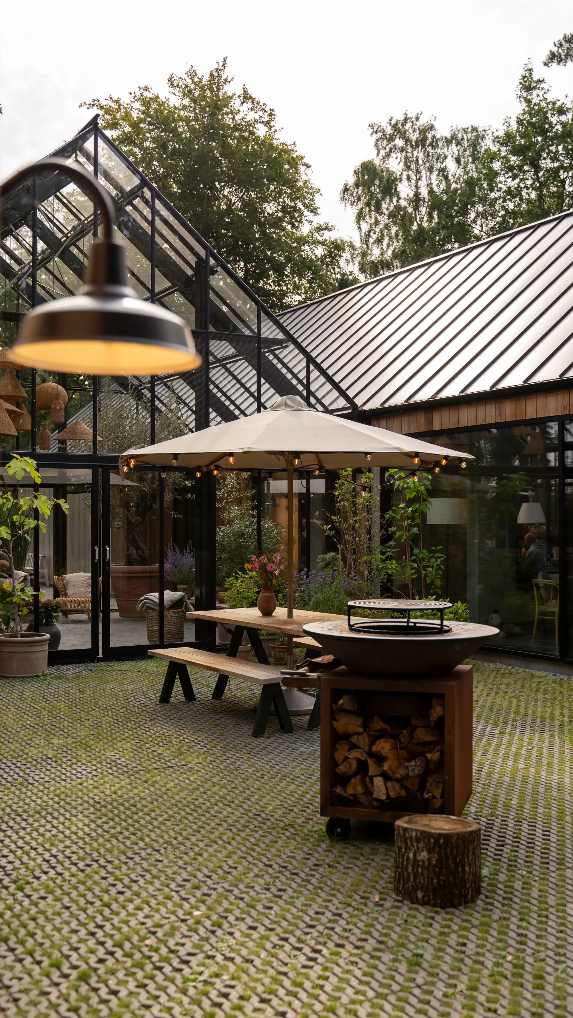 Hyggelig gårdhave med glasdrivhus, langtræsbord under parasol og udepejs med brænde.