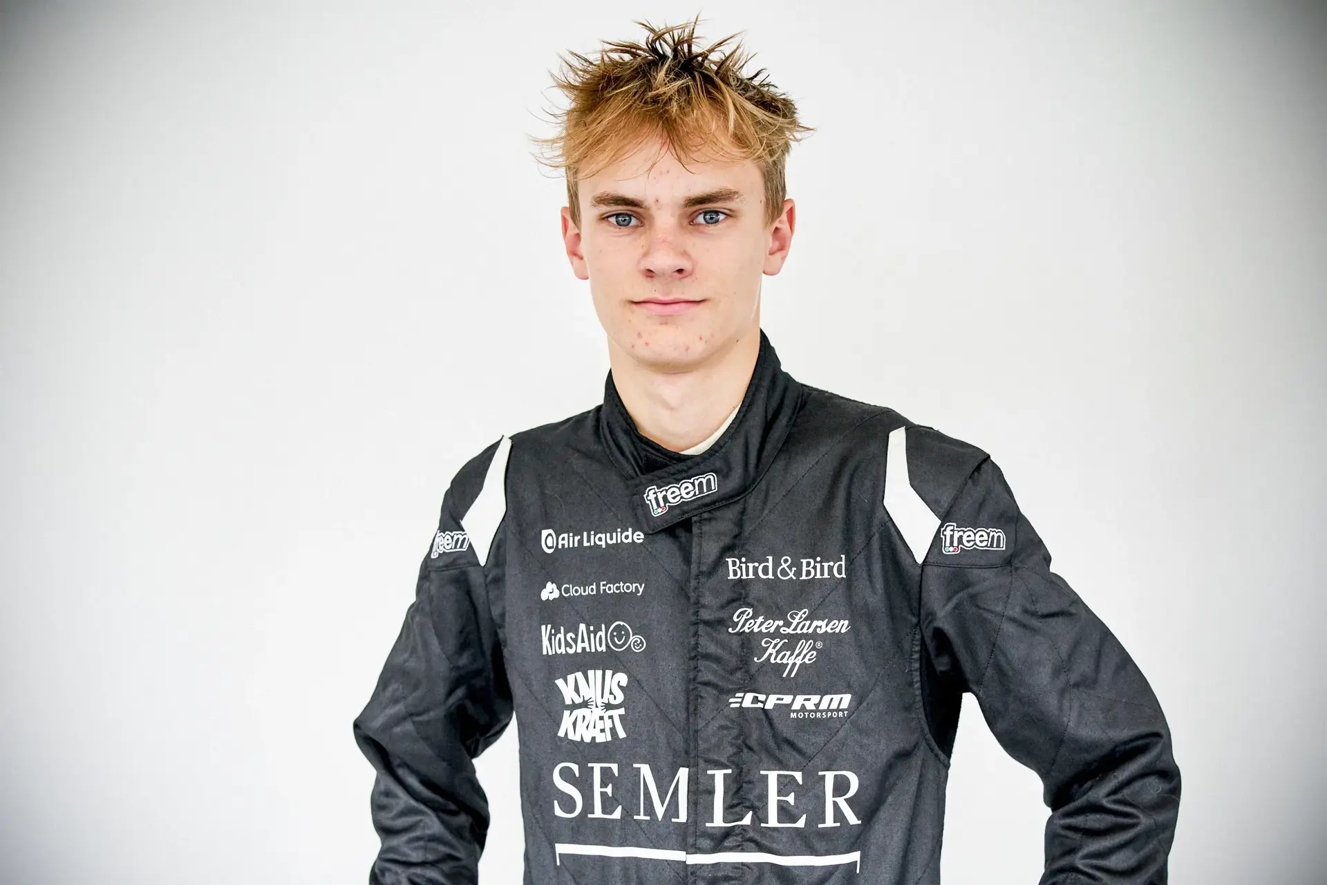 Racemagnetic ifører sig en sort køredragt med flere sponsorlogoer foran en neutral baggrund.