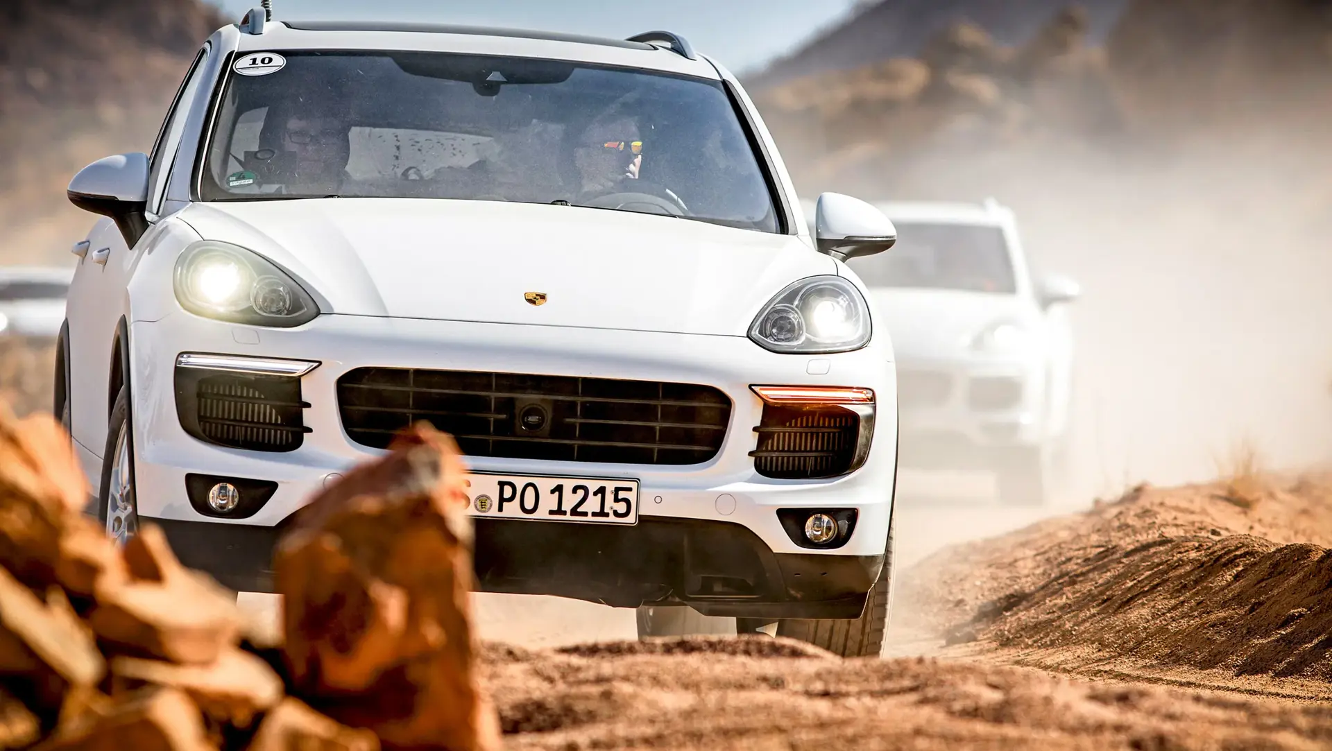 En hvid Porsche Cayenne S kører i høj fart på en støvet grusvej gennem et øde, klippefyldt landskab.