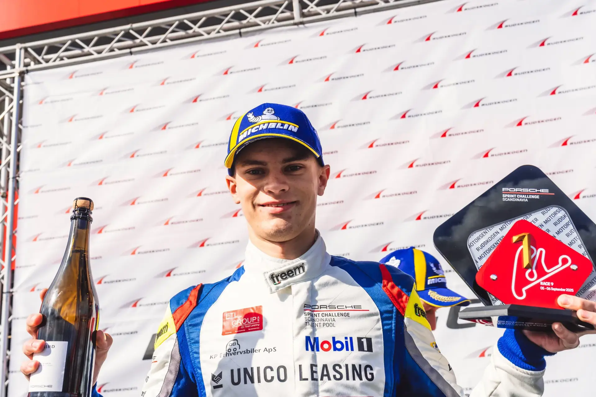 Racerkører på sejrspodiet holder en champagnemagnum og et Porsche Sprint Challenge-vindertrofæ.