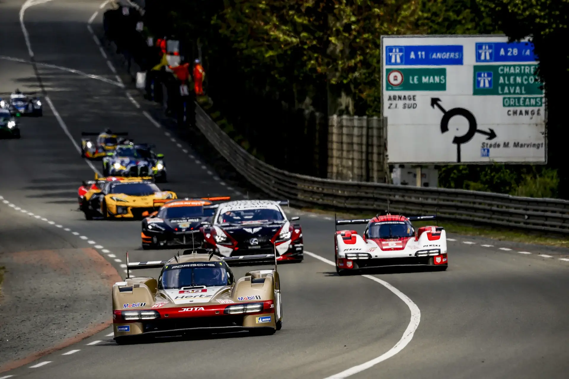 Sportsvogne i høj fart racer ned ad en vej under 24-timersløbet på Le Mans, med vejskilte synlige i baggrunden.