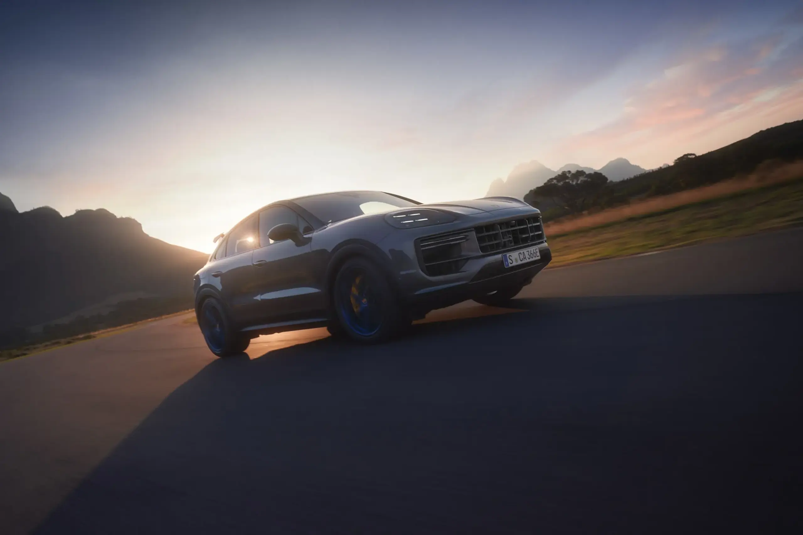 En mørkegrå Porsche Cayenne Coupé kører i høj fart på en vej ved solnedgang med bjerge i baggrunden.