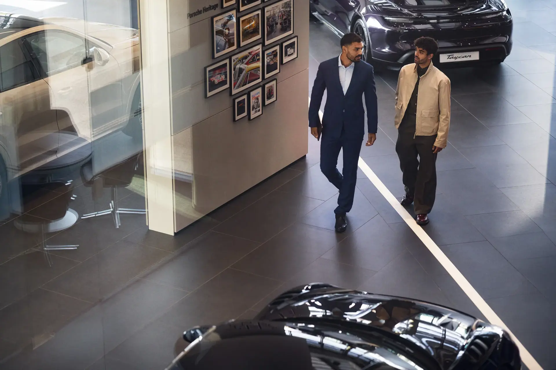 To mænd går og taler sammen i et Porsche-bilhus omgivet af udstillede biler.